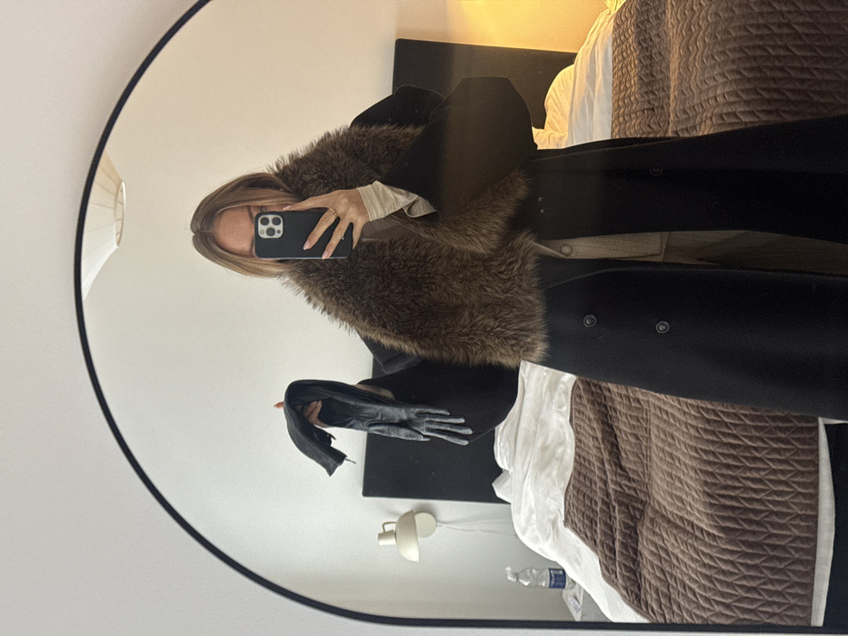 A mirror selfie captures an individual, a Caucasian female, standing in a bedroom in a residence in Unknown, Unknown. The subject, partially obscured by a dark-cased smartphone held in her right hand, wears a long black double-breasted coat featuring a prominent brown faux fur collar, over a light-colored long-sleeved top and beige trousers. She holds a pair of black leather gloves in her extended left hand. The reflection reveals a minimalist bedroom setting: an unmade bed with white sheets and a brown quilted blanket, a dark, simple headboard, a cream-colored wall-mounted lamp to the left, and a clear plastic water bottle on a bedside surface. A white circular ceiling lamp fixture is visible at the top center.