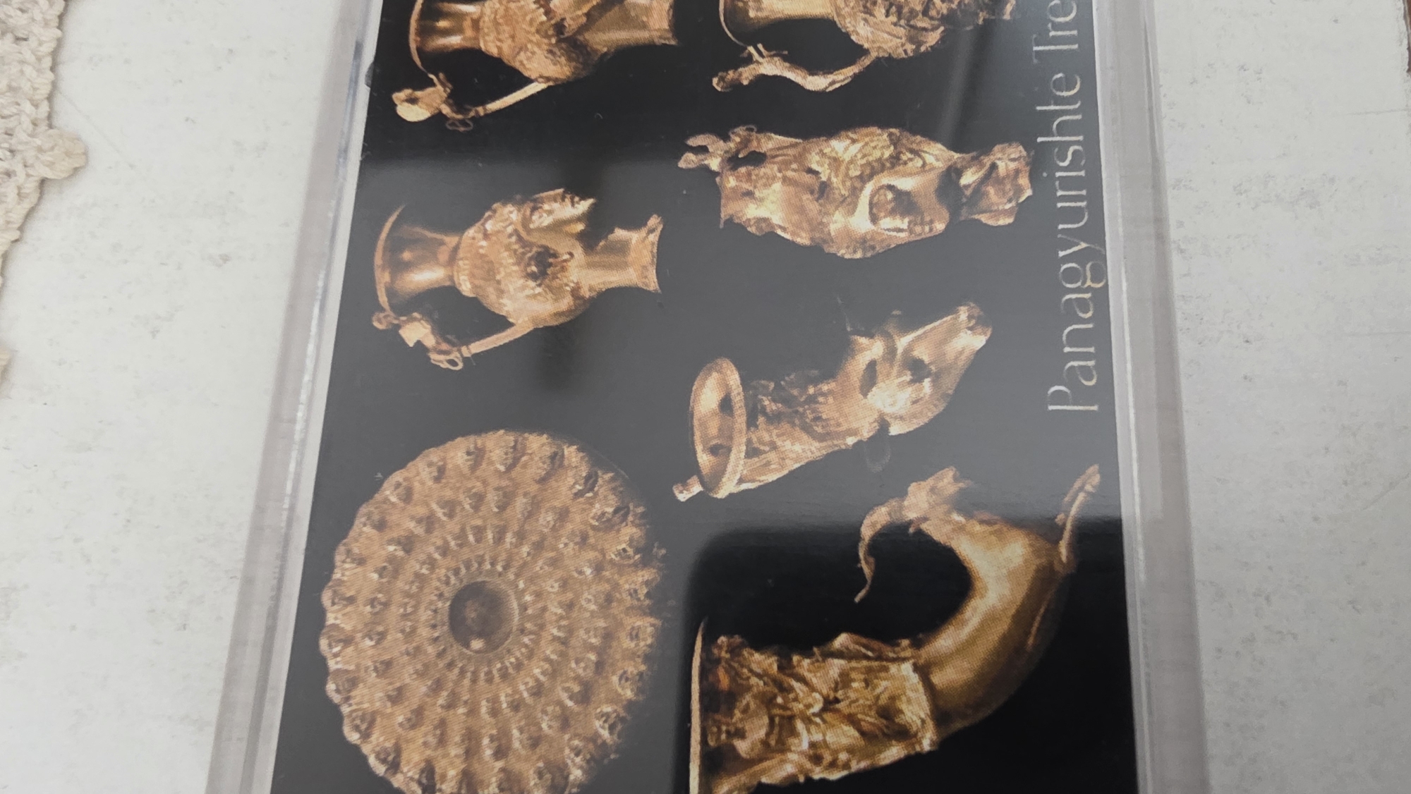 The image shows a postcard of the Panagyurishte Treasure, a Thracian gold hoard discovered in Panagyurishte, Bulgaria. The inscription "Panagyurishte Tre" is visible on the right side. The objects depicted appear to be various golden vessels and artifacts. No people, actions, or other environmental details are present in the image. The location information is inferred to be in Blagoevgrad, Bulgaria as the postcard is from there.