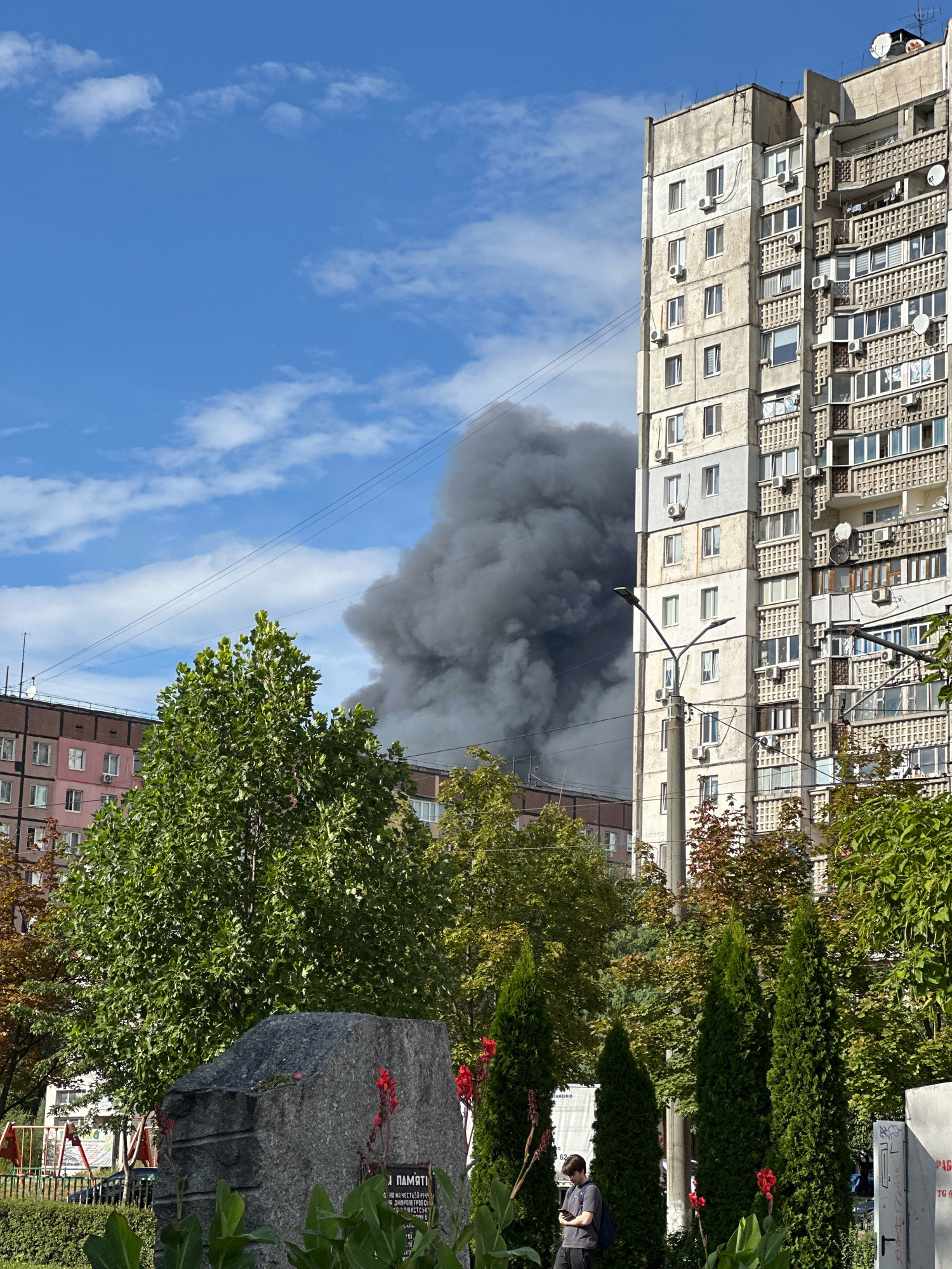 In Dnipro, Ukraine, on a bright, partly cloudy day, a large plume of dark smoke billows upwards, obscuring the sky behind a row of trees and apartment buildings. The smoke appears to originate from a fire in the distance, likely within a residential area. In the foreground, a large, rough-hewn granite monument with inscriptions stands amidst green foliage and red flowers. To the right of the monument, a young person wearing a dark shirt and backpack is looking down at their phone. Behind the trees, several multi-story apartment buildings, typical of Soviet-era architecture, are visible, with the tall, concrete structure on the right featuring numerous balconies and windows. Power lines are strung across the sky in several places. The scene suggests a moment of disruption and potential crisis amidst a normal urban environment.