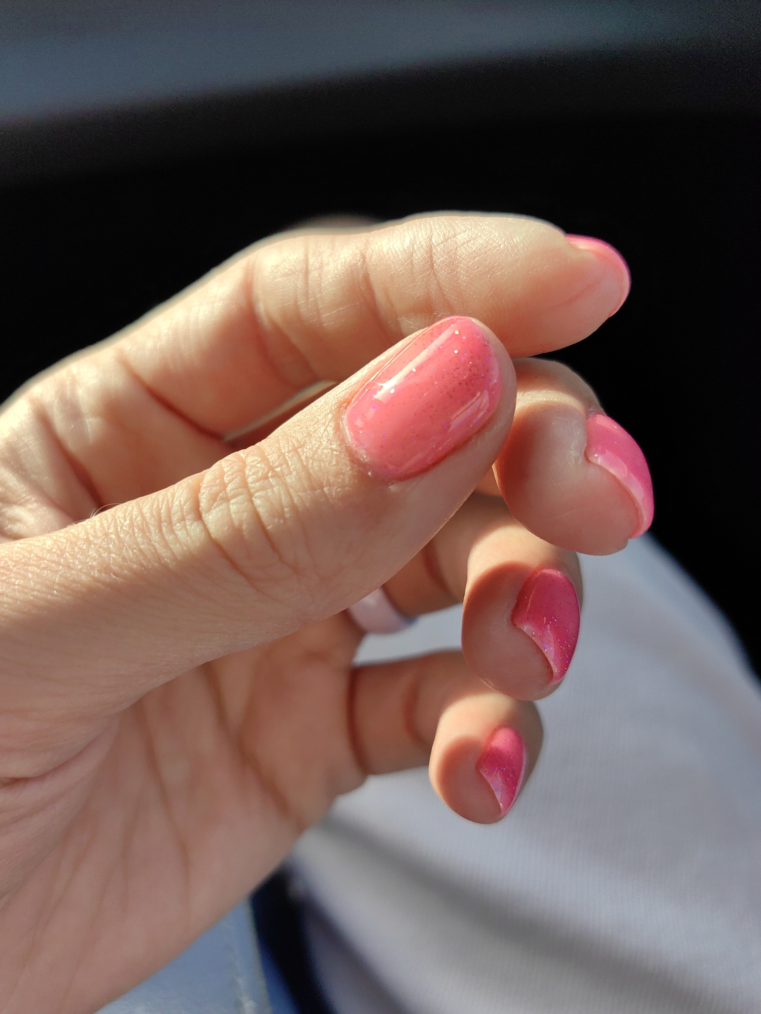 The image shows a close-up of a person's hand with bright pink, glittery nail polish on their fingernails. The hand is positioned against a dark background, with some light creating shadows on the fingers. The skin appears to have visible texture and creases. The background consists of a dark area, possibly a shadow or a dark object, and a lighter area with a subtle texture, which could be fabric. The specific location in Путчынскі сельскі Савет, Belarus is not discernible from the visual information provided in the image.