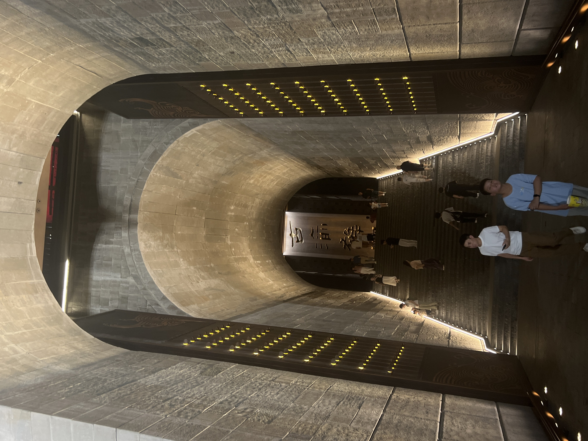 The image shows a grand stone staircase leading up into a large, vaulted tunnel-like structure. The walls are made of rough-hewn stone blocks, and vertical panels with rows of small, bright yellow lights line the sides of the staircase. At the top of the visible staircase, a large banner with Chinese characters is illuminated. Several people are ascending the stairs, walking in various directions. In the foreground, two young males are walking towards the camera; one is wearing a white t-shirt and brown pants, holding a mobile phone, and the other is wearing a light blue t-shirt and grey pants, holding a bucket of popcorn. The lighting is dim and dramatic, with the yellow lights and illumination from above creating stark shadows. The architectural style suggests a historical or ceremonial site. This scene is located at the Yangtze River Cableway in Yuzhong District, Chongqing, China.