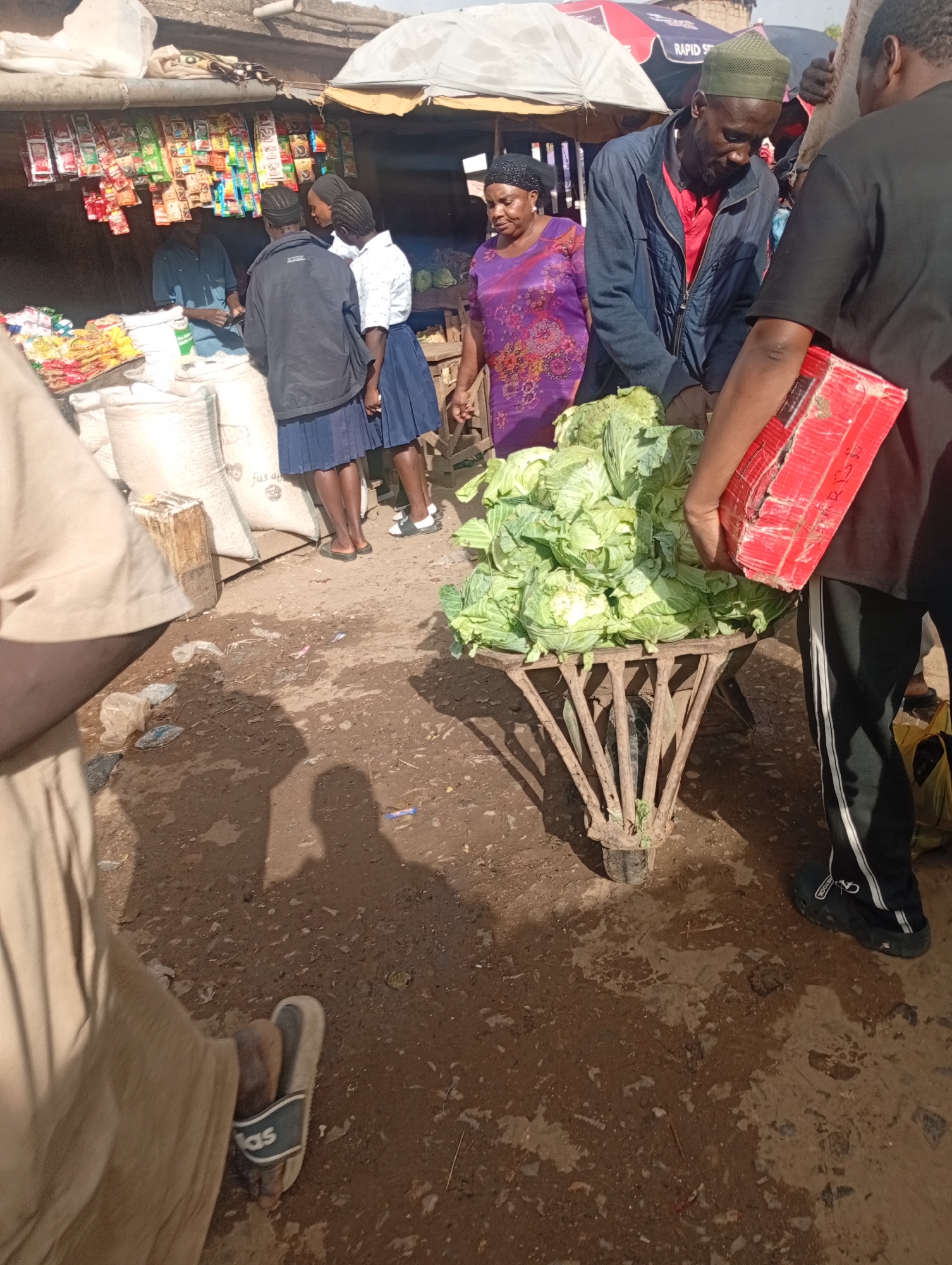 In a bustling outdoor market in Rafin Par, Nigeria, several individuals are engaged in commerce. A man wearing a green cap and blue jacket is focused on a large pile of cabbages loaded onto a metal cart. Beside him, a woman in a vibrant purple dress stands, observing. The cart is being attended to by another person, partially visible, holding a red cardboard box.
In the background, a stall displays an array of colorful packaged goods, suggesting a variety of food items or products. Two young women, possibly students given their matching blue skirts and white shirts, are interacting near the stall. Large sacks, likely containing grains or other staples, are stacked behind them. The ground is a mix of dirt and scattered debris, indicative of a busy marketplace. The scene appears to be during daylight hours, with the sun casting shadows on the ground. The overall atmosphere suggests a typical day of trading and social interaction within the community.