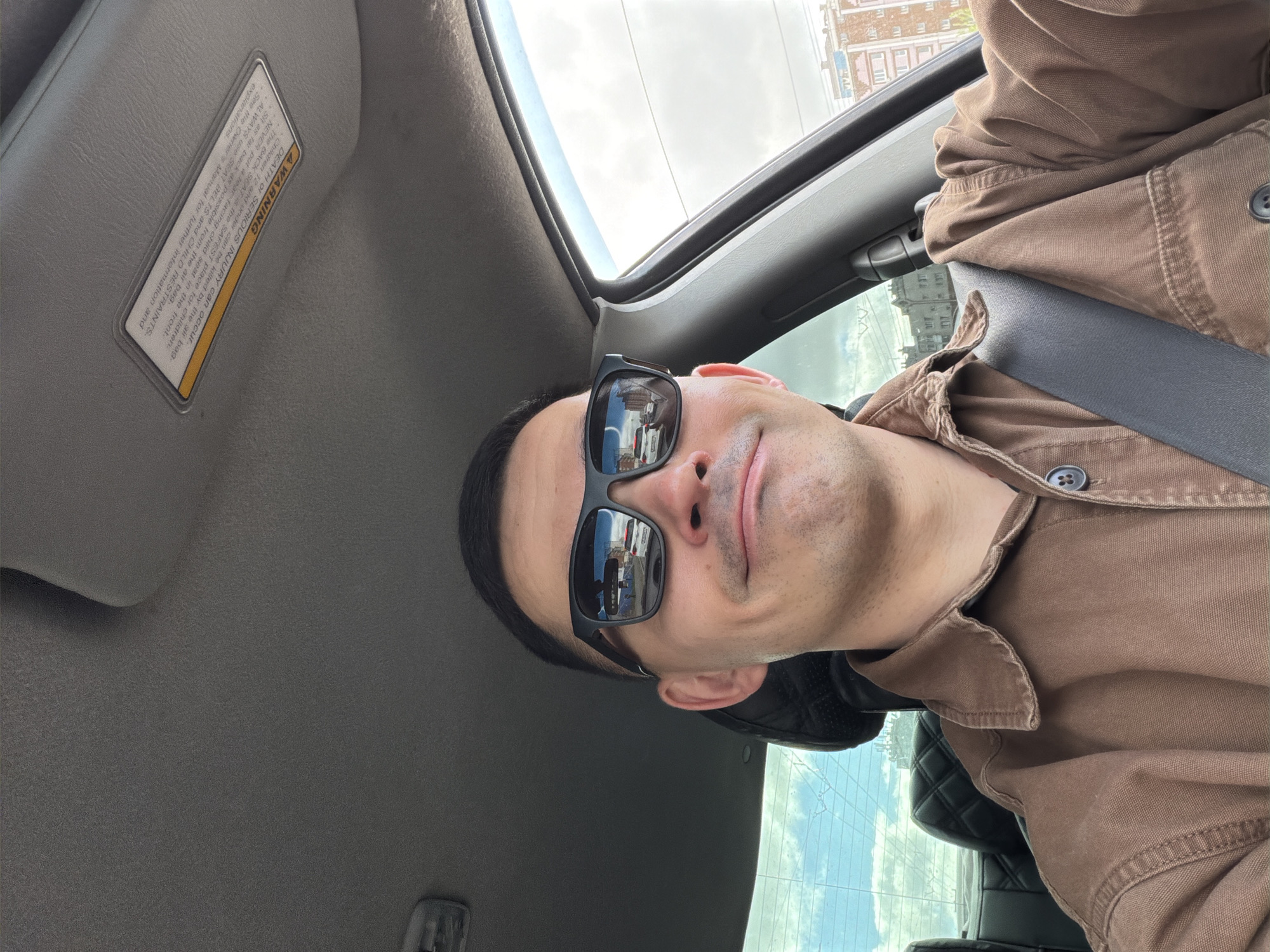 A man wearing sunglasses and a brown button-down shirt is in the driver's seat of a car. His seatbelt is fastened. A building is visible in the reflection of his sunglasses. The visor on the driver's side of the car is down, with a warning label visible. The car appears to have a sunroof. The location is implied to be Izhevsk, Russia due to the reflection in the sunglasses.
