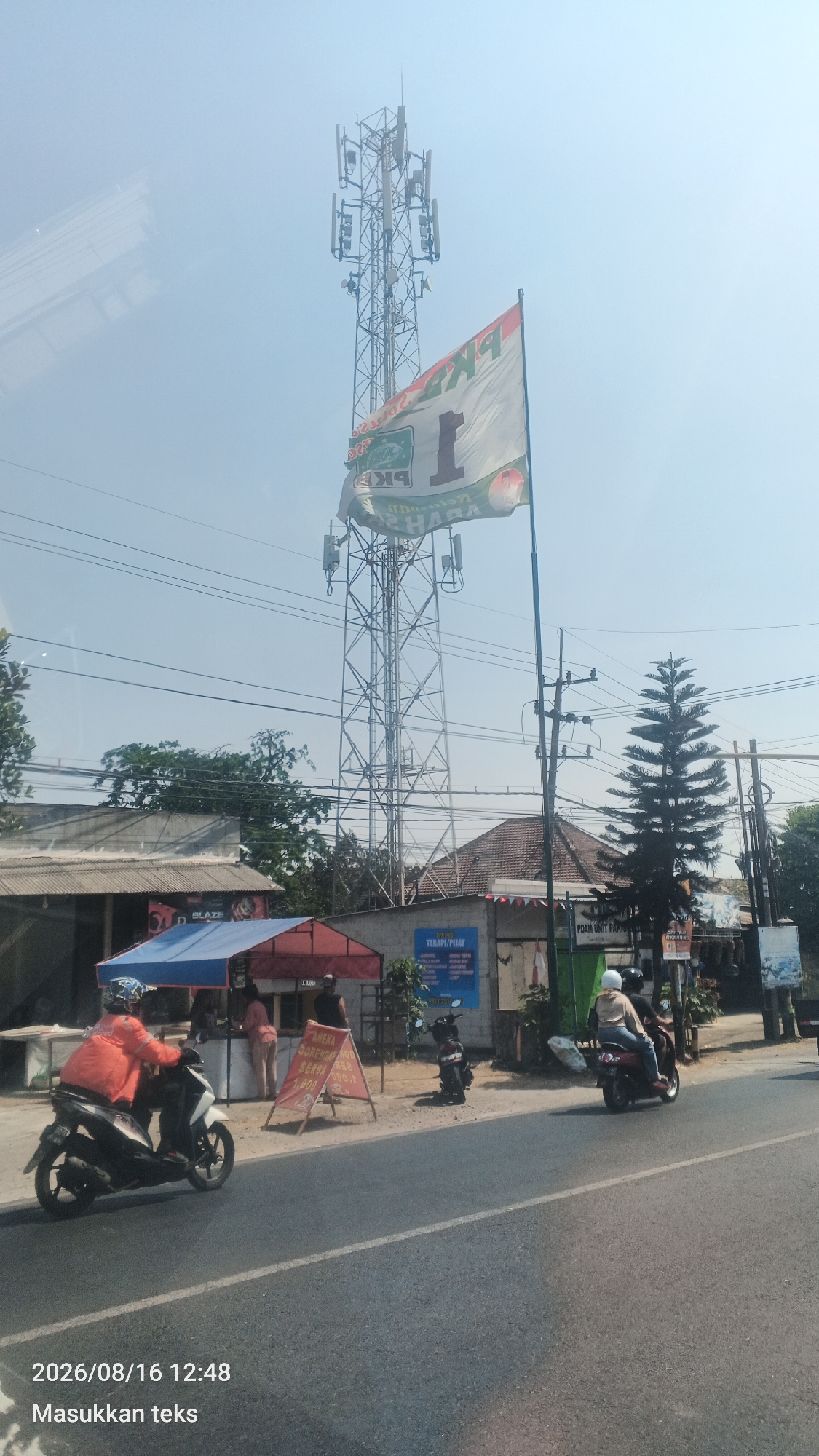 An outdoor scene on a sunny day shows a cell tower in Pakis, Indonesia. The tower is tall and metallic, with multiple antennas attached to it. A large white flag with a green and red design, and the number "1" prominently displayed, is attached to a flagpole near the tower and billows in the wind.
In the foreground, a paved road runs horizontally across the image. On the right side of the road, there is a sidewalk with several buildings and structures. One building has a blue and red awning over a small stall where two people are standing. Further down, another building has a sign that reads "TERAPI PIJAT." A pine tree stands to the right of these structures.
Several motorcycles are present. A person wearing an orange jacket is riding a white motorcycle on the road, moving from left to right. Another motorcycle is parked on the sidewalk. Two people are riding on a dark-colored motorcycle moving from right to left on the road.
The background consists of a clear blue sky with some faint reflections on the left side, possibly from a window. The overall environment suggests a roadside scene in a built-up area. The date stamp "2026/08/16 12:48" is visible in the lower left corner of the image.