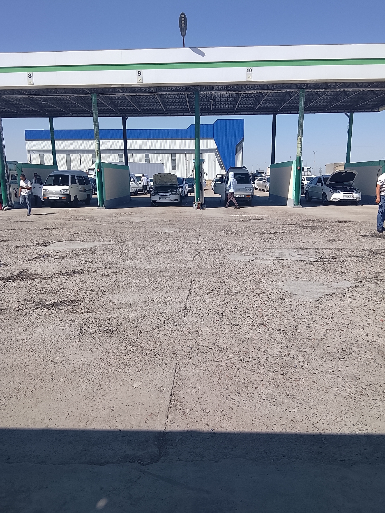 The image shows a car service station or parking area under a covered structure. The structure has pillars and a white roof with green trim. Numbers "8", "9", and "10" are visible on the roof, indicating bays or stalls. Several vehicles are parked or being serviced in the bays. In bay 8, a white van is parked, and a man in a white shirt and blue jeans is walking away from it. In bay 9, another white van is visible. In bay 10, a white car with its hood open is parked, and a person in a white shirt and dark pants is standing near it. Another white car is parked behind it. The ground is paved with rough asphalt. In the background, there is a large building with blue and white sections, and a clear blue sky. The overall scene suggests a commercial area, possibly a marketplace or service center for vehicles. The presence of specific stall numbers and visible car servicing activities points towards a functional establishment. The presence of the landmark building in the background likely helps in identifying the specific location within Syrdarya, Uzbekistan.