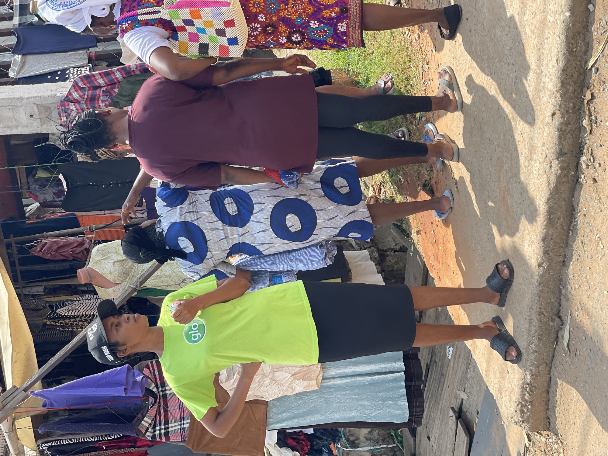 This image shows four women standing at an outdoor market stall. The woman in the foreground is wearing a bright green t-shirt with a "glo" logo and a black pencil skirt, and a black baseball cap. She is looking to her left and has her right hand on her hip. To her right, a woman in a blue and white patterned dress is looking forward and holding a red and blue folded cloth. Behind her, a woman in a maroon t-shirt and black leggings is facing away from the camera, with her hands behind her back. To the right of the woman in maroon, another woman is wearing a white top and a brightly colored patterned skirt, holding a woven bag on her right shoulder. The market stall behind them is filled with hanging clothes. The ground is a mixture of dirt, concrete, and wooden planks. Sunlight casts shadows on the ground.