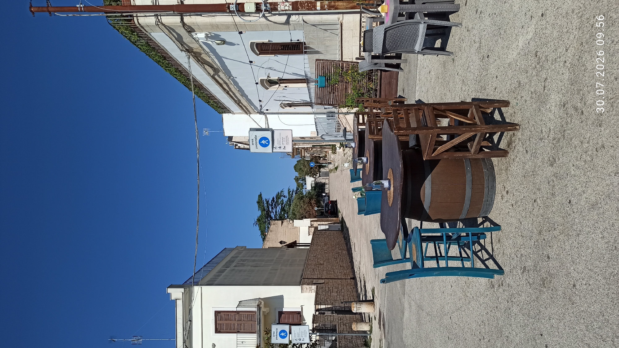 The image depicts an outdoor seating area of a cafe or restaurant on a sunny day. In the foreground, several tables and chairs are arranged on a paved surface. Two tables are prominent: one is a round table with a dark brown top, supported by a large wooden barrel base, and surrounded by three blue bar stools. Adjacent to it is another table with a similar barrel base, accompanied by wooden chairs. Further back, other tables and chairs, including grey plastic chairs, are visible. The setting appears to be a street or a public square, with buildings lining both sides.
On the left side of the street, a white building with a balcony and brown shutters is prominent. A stone wall runs along the front of this property. On the right side, another building with light-colored facade and windows is visible, along with planters and decorative string lights.
Two blue and white pedestrian zone signs are posted. The sign on the left features a pictogram of a person walking and the text "AREA PEDONALE." The sign on the right is similar, with the same pictogram and text, indicating a pedestrian area.
In the background, the street recedes, with more buildings and trees visible. A car is parked on the street in the distance. The sky is a clear, deep blue.
The timestamp "30.07.2026 09:56" is overlaid in the bottom right corner of the image, indicating the date and time the photo was taken.
The location is identified as Terlizzi, Italy. This specific scene is likely in the historic center or a pedestrianized area of the town, given the pedestrian zone signage and the architectural style of the buildings.