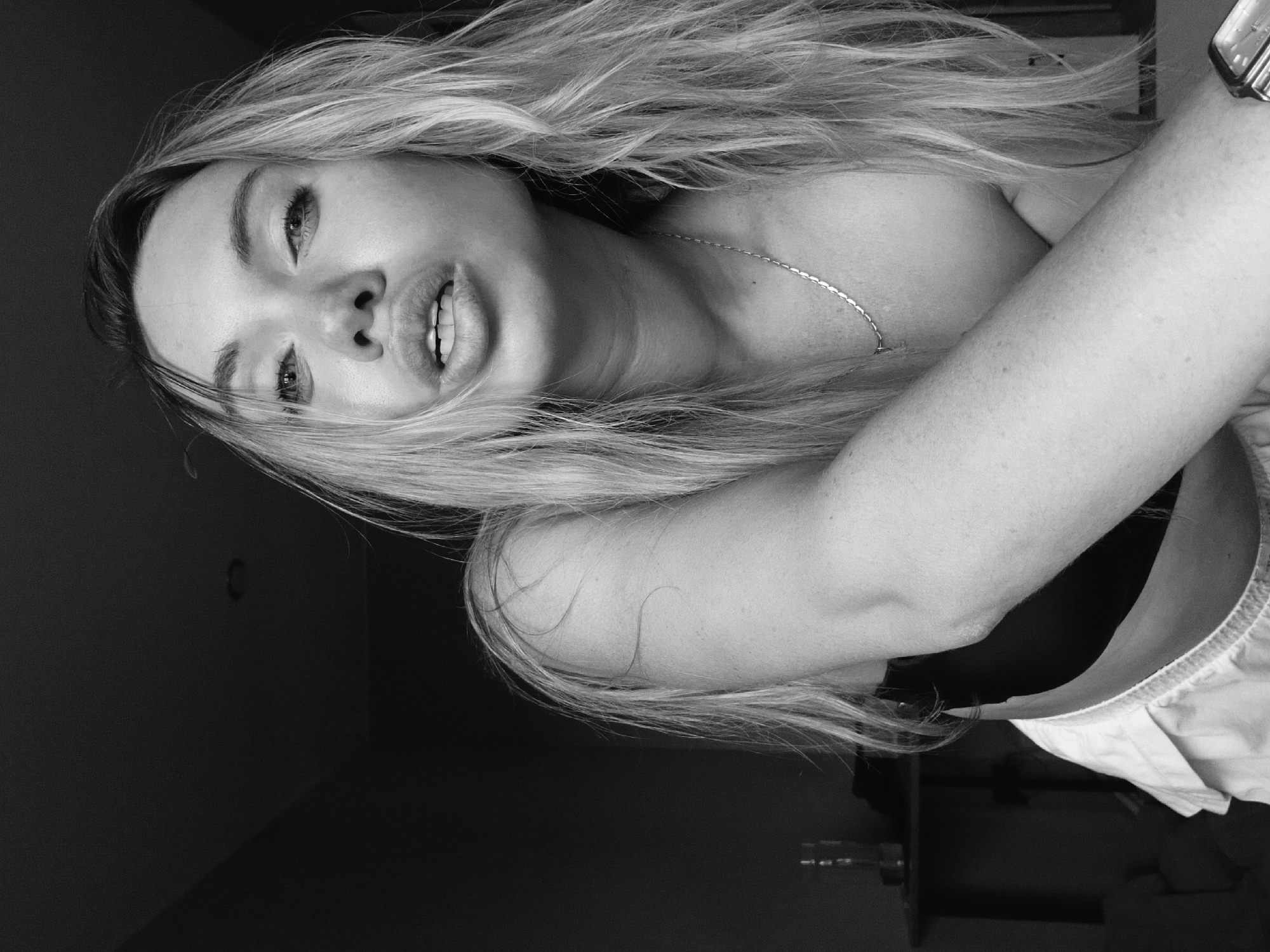 A monochrome, low-angle portrait captures a woman with light-colored hair, positioned at an angle relative to the camera. She is wearing a delicate necklace and a garment with light-colored fabric visible at the bottom edge. Her expression is captured with her lips slightly parted. The background is predominantly dark, with some indistinct objects. There is no indication in the image that would indicate that this image was taken in Votkinsk, Russia.