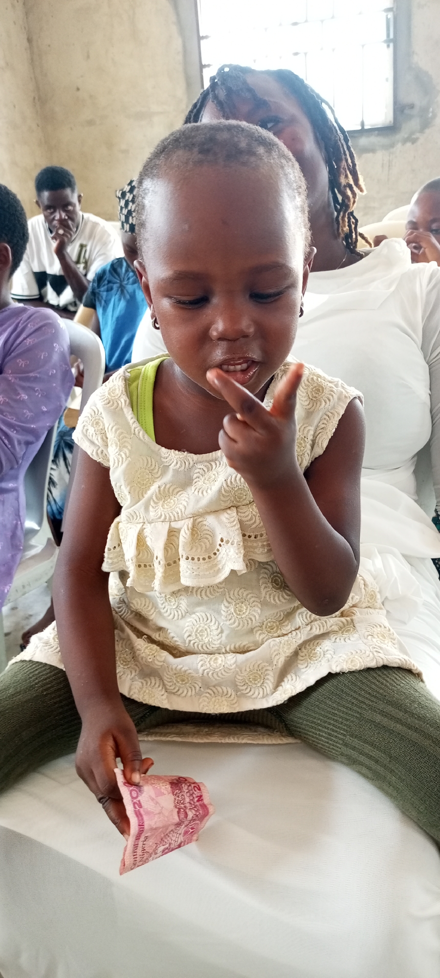 A young girl sits on a white surface, possibly a chair, holding a folded pink banknote in her left hand and gesturing with her right hand towards her mouth. She wears a cream-colored dress with a ruffled design and green leggings. Behind her sits an adult wearing a white top, with dreadlocks visible. Other people are visible in the background, including a man in a white and black striped shirt. The location is unknown.