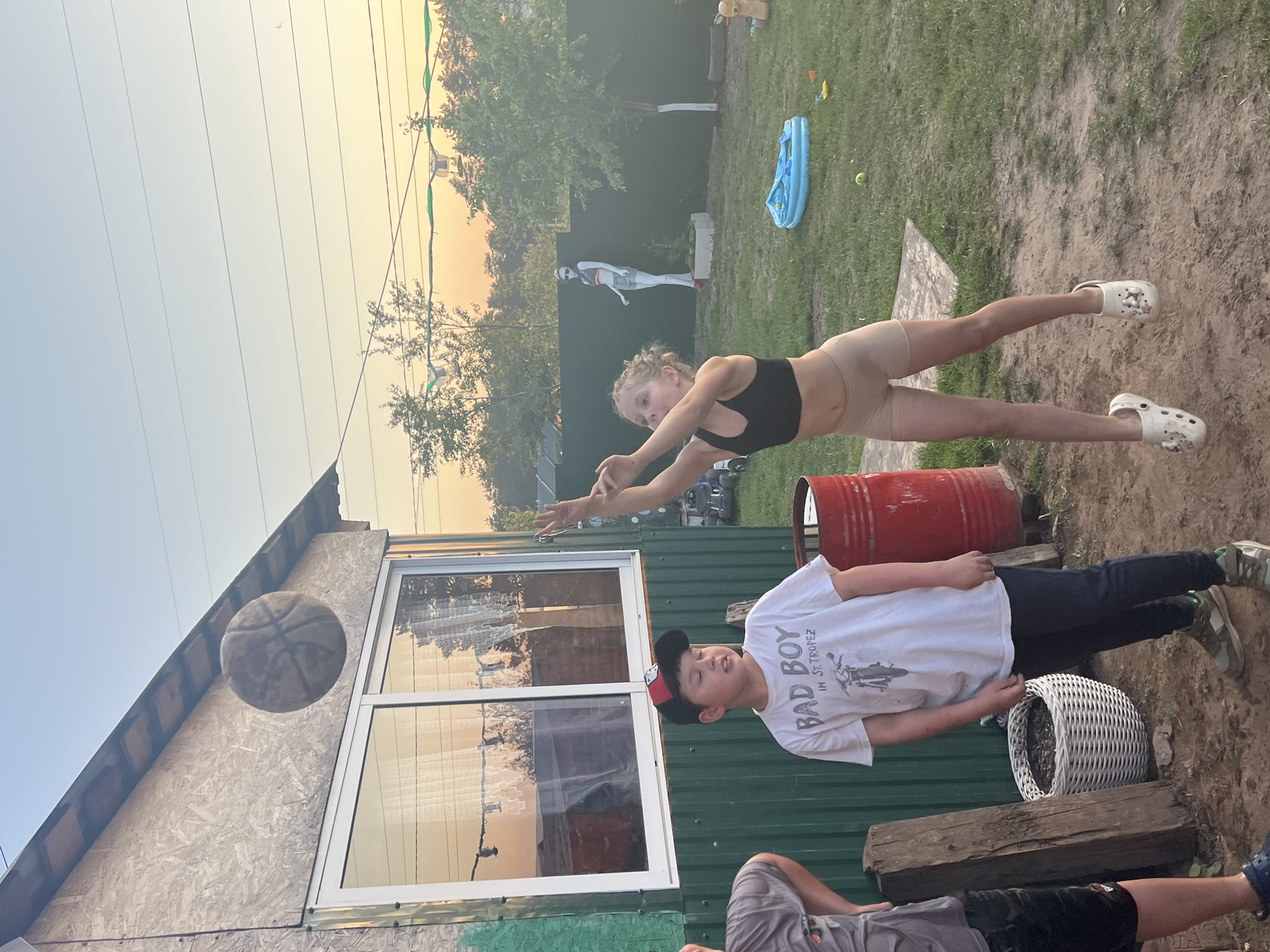 Location: Chaikovsky, Russia.
Exterior shot. A woman in a black sports bra and beige shorts is airborne, arms outstretched. A boy in a white t-shirt with a black and red baseball cap is lying on a green corrugated metal surface. A basketball is in the air near the corner of a building. Visible are a red barrel, a white wicker basket with dark contents, and two wooden logs. Faintly visible in the background is a white figure by a fence and some greenery. The sky is clear.