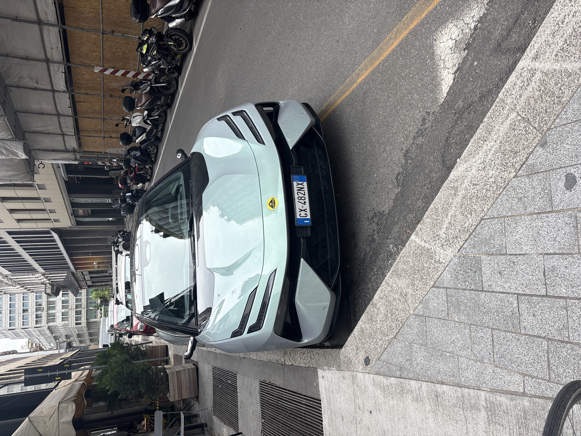 The image shows a pale green Lotus Emira parked on a city street in Milan, Italy. The car is positioned next to a curb, and the license plate number is GX-482NX. There are rows of parked motorcycles alongside a building under construction to the left of the car. The street has a yellow line, and other vehicles are parked further down the street. The background features buildings, indicating an urban setting. There are also trees in wooden boxes and a bicycle wheel visible. The photo appears to be taken during the day.