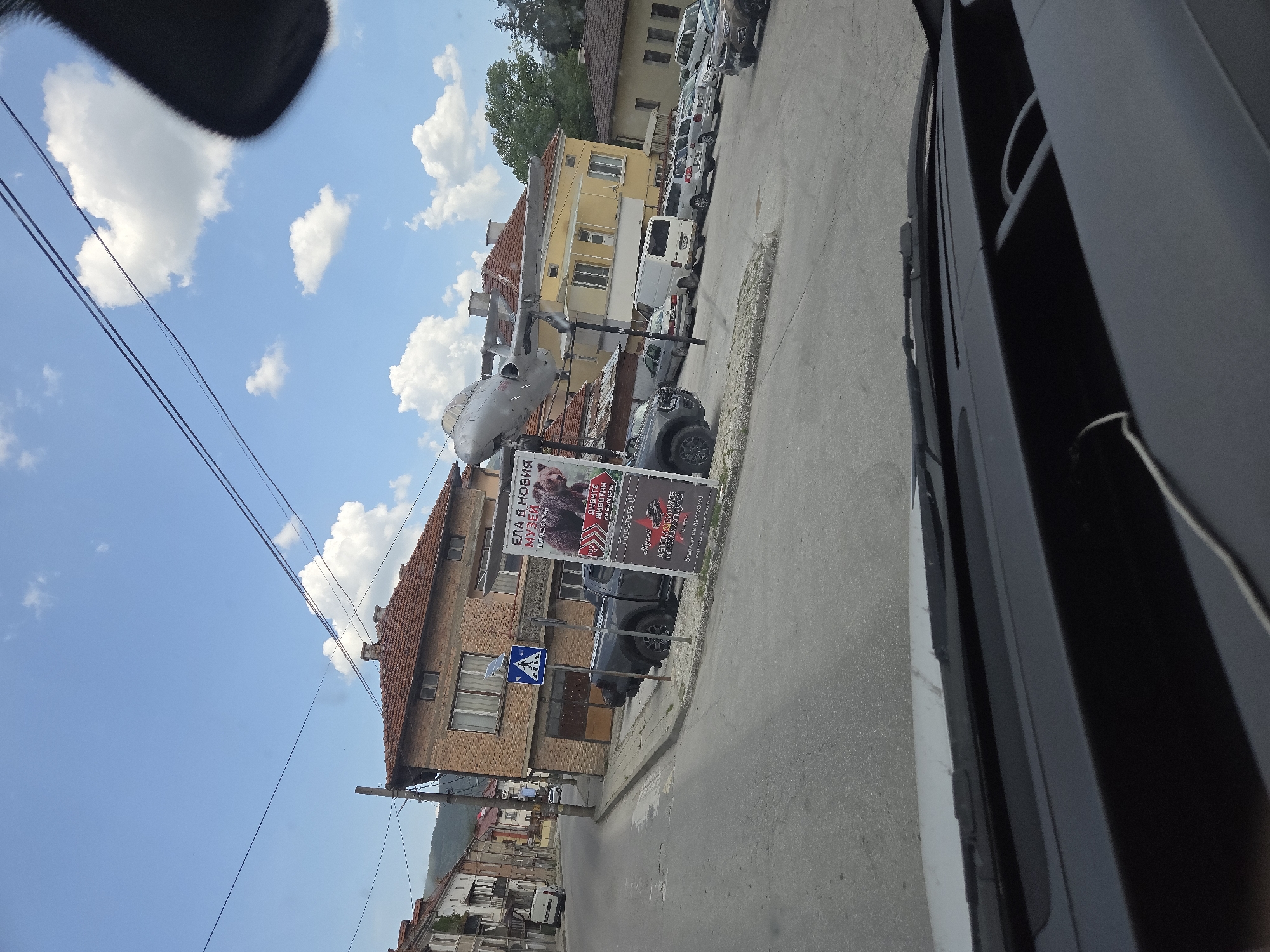 Here is a detailed description of the image:
**Setting:**
The image appears to be taken from inside a vehicle, likely a car or van, parked or driving along a street in a town or city. The weather is sunny with blue skies and scattered clouds.
**Objects and Subjects:**
* **Buildings:** On the side of the street, there are several buildings, mostly two to three stories high, with red tile roofs and brick or painted facades. Their architectural style seems typical of European or Eastern European towns.
* **Street and Sidewalk:** The street is paved and appears to be of moderate width. There is a sidewalk along the buildings.
* **Cars:** A number of cars are parked along the side of the street, suggesting a residential or commercial area.
* **Sign:** There is a large advertisement sign erected next to the sidewalk. The sign is partly visible and contains text in a Cyrillic alphabet, indicating a possible location in Bulgaria. The text on the sign appears to be promoting a museum exhibit, "ЕЛА В НОВИЯ МУЗЕЙ" (Come to the new museum).
* **Airplane Propeller Monument**: A gray airplane with two propellers is placed on the side of the street.
**Actions and Interactions:**
* The image is a static snapshot, so there are no actions directly visible. However, the presence of parked cars and buildings implies a residential and commercial environment where people live, work, and move around.
* The person taking the photo is inside a vehicle and likely either driving or observing the scene.
**Overall Impression:**
The image captures a common urban scene in a European town or city during a bright day. The presence of the sign indicates a specific place, possibly a business or tourist attraction.
**Location:**
Based on the Cyrillic text on the sign, the location could be in Bulgaria.