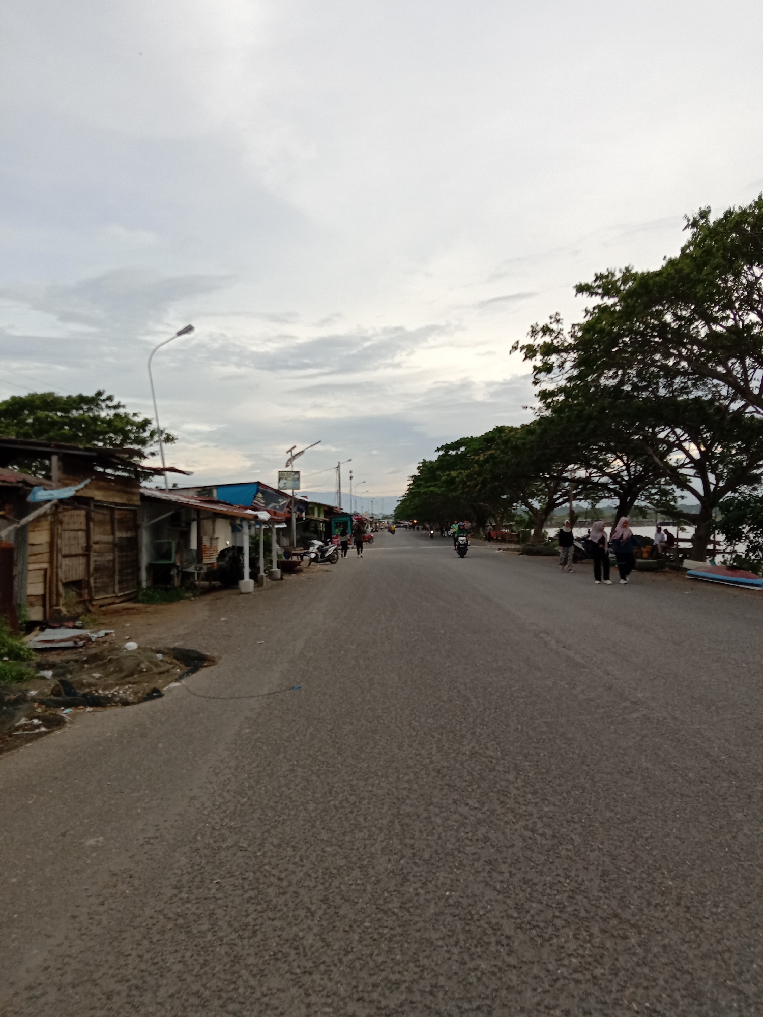The image depicts a long, straight asphalt road in Lhokseumawe, Indonesia. Along the left side of the road are makeshift wooden structures and shacks. On the right side, mature trees line the road. Several people, some wearing headscarves, are walking along the right side of the road and some are by the water. Motorbikes are traveling on the road. Overhead, the sky is overcast with gray and white clouds. Lampposts are visible along the road.