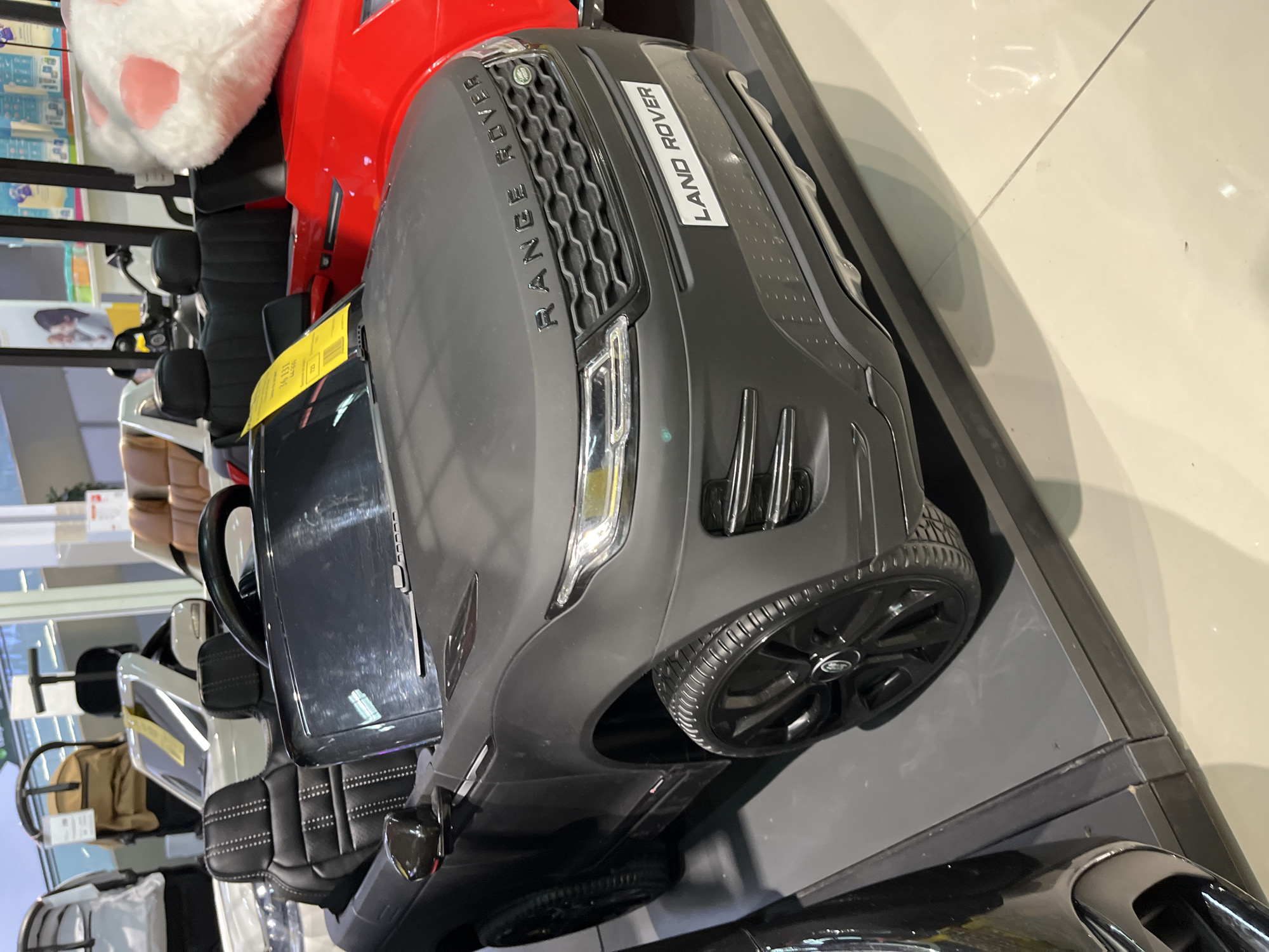 This image depicts an indoor retail environment located in Sochi, Russia, showcasing children's ride-on electric vehicles and other child-related merchandise.
The central subject is a matte black, two-seat, child's ride-on electric vehicle, designed as a replica of a Range Rover Evoque. The words "RANGE ROVER" are embossed in black above the front grille, which features a honeycomb pattern and a small Land Rover badge. Below the grille, a white rectangular plate reads "LAND ROVER." The vehicle is equipped with black multi-spoke wheels fitted with rubber-textured tires and features illuminated or reflective headlights. A yellow price or information tag is affixed to the passenger side of the windshield. The interior reveals black seats with detailed white stitching patterns.
To the immediate right of the black vehicle, a portion of a second, red, ride-on electric vehicle is visible, also displayed on the same platform. A large, fluffy white plush object with pink pads, resembling a cartoon animal's paw or ear, rests on the hood of the red vehicle.
Both vehicles are positioned on a dark grey, multi-tiered display platform. The floor of the establishment is covered in light-colored (likely beige or off-white) tiles, exhibiting reflections from the overhead artificial lighting.
In the background to the left, various infant and child care items are visible, including a grey stroller-like object and a tan child car seat or stroller, both presented on individual display stands. Further in the background to the right, retail shelving stocked with packaged products and an advertising poster depicting an adult with a child are discernible, reinforcing the commercial retail setting.