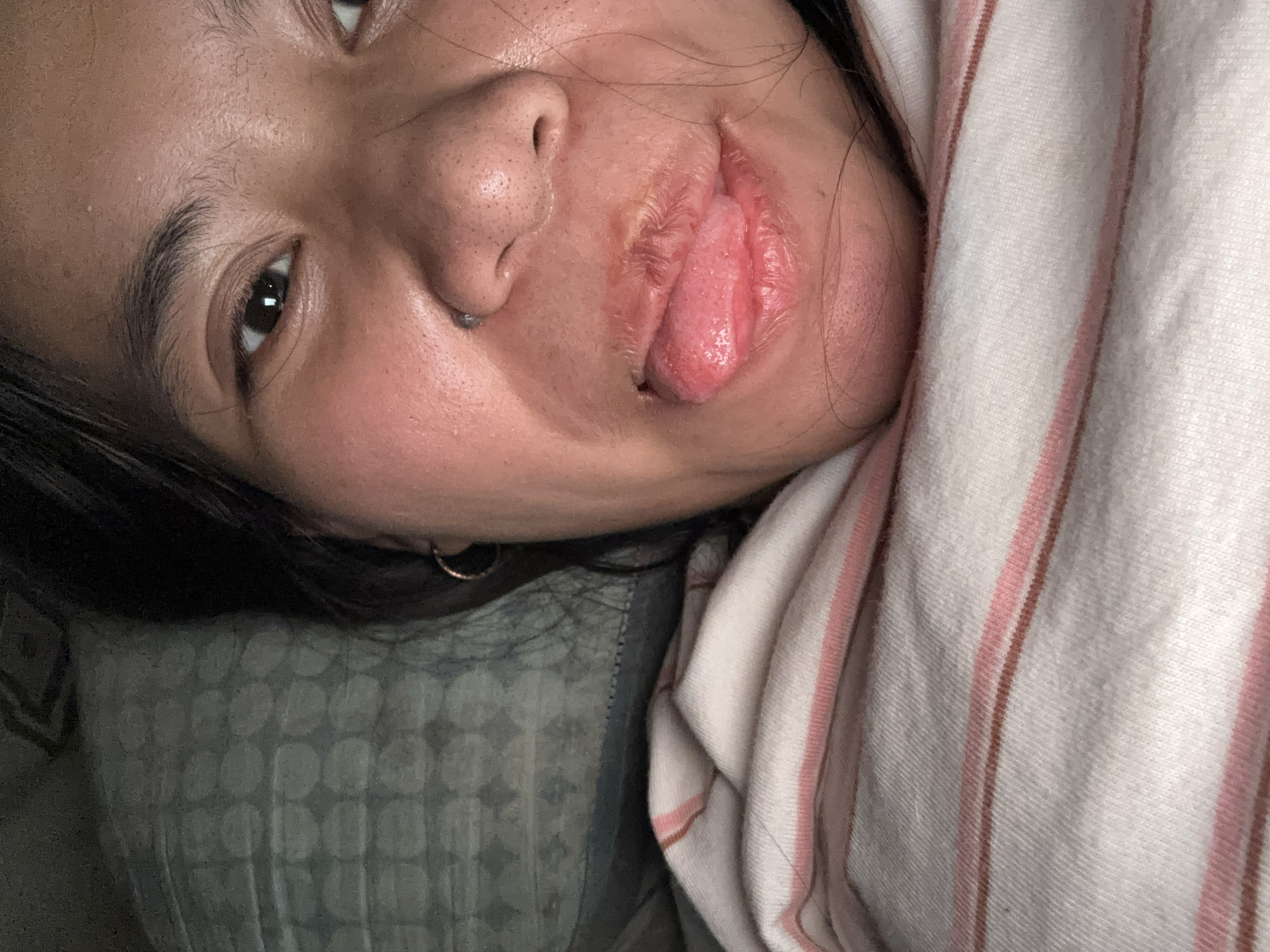 The image is a close-up selfie of a person lying down, with their face filling most of the frame. The person has dark hair, brown eyes, and a nose ring. Their lips are slightly parted, and their tongue is sticking out. The person is wearing a striped shirt. The background appears to be a pillow with a circular pattern. The image is taken indoors, likely in a bedroom. There are no identifying landmarks or details that would allow for the determination of its location in East Jakarta, Indonesia.
