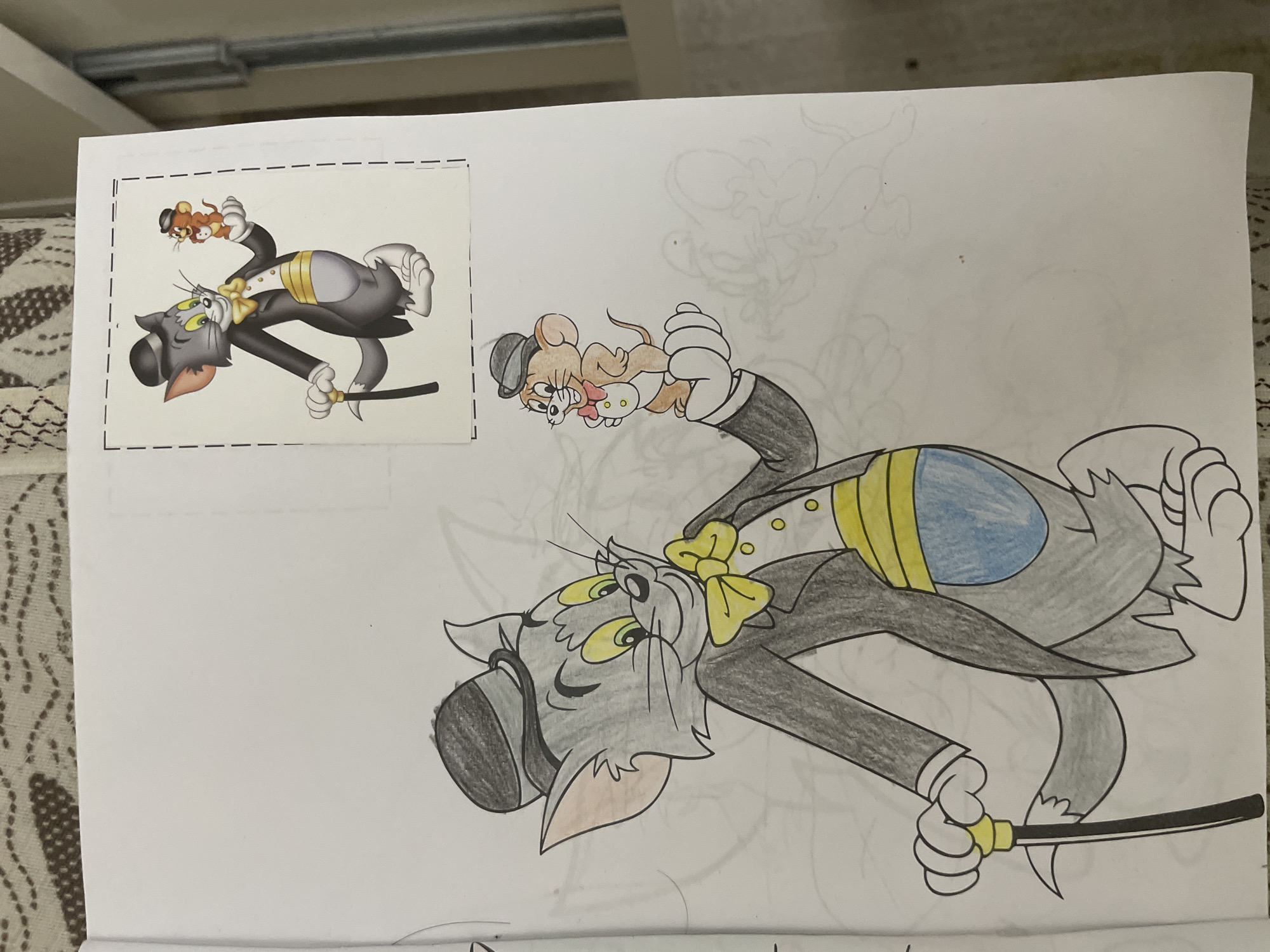 The image features a partially colored drawing of the cartoon characters Tom and Jerry on a sheet of white paper, likely a coloring page or a traced image. In the upper left corner, a smaller, fully colored reference image of the same scene is pasted, outlined by dashed lines.
Tom, the grey cat, is depicted lying on his back, dressed in a formal grey tuxedo with a yellow bowtie and a grey top hat. His cummerbund is striped yellow and blue. He holds a black cane with a yellow handle in his right paw. His eyes are green, and he has a surprised or curious expression, looking upwards.
Jerry, the brown mouse, is perched on Tom's stomach, also looking in the same upward direction. He wears a small grey bowler hat and a red bowtie, with white cuffs visible on his tiny suit sleeves.
The main drawing is partly colored, with Tom's tuxedo, bowtie, cummerbund, and Tom and Jerry's facial features and some fur areas filled in. However, parts of Jerry's body and other details remain as black outlines. Faint pencil sketches of other cartoon figures or elements are visible in the background of the main drawing, indicating an unfinished sketch or draft. The paper is placed on a surface, with a patterned brown and white textile visible to the left. In the background of the original photo, a white ceiling with parallel fixtures can be seen. No explicit text is visible in the image.