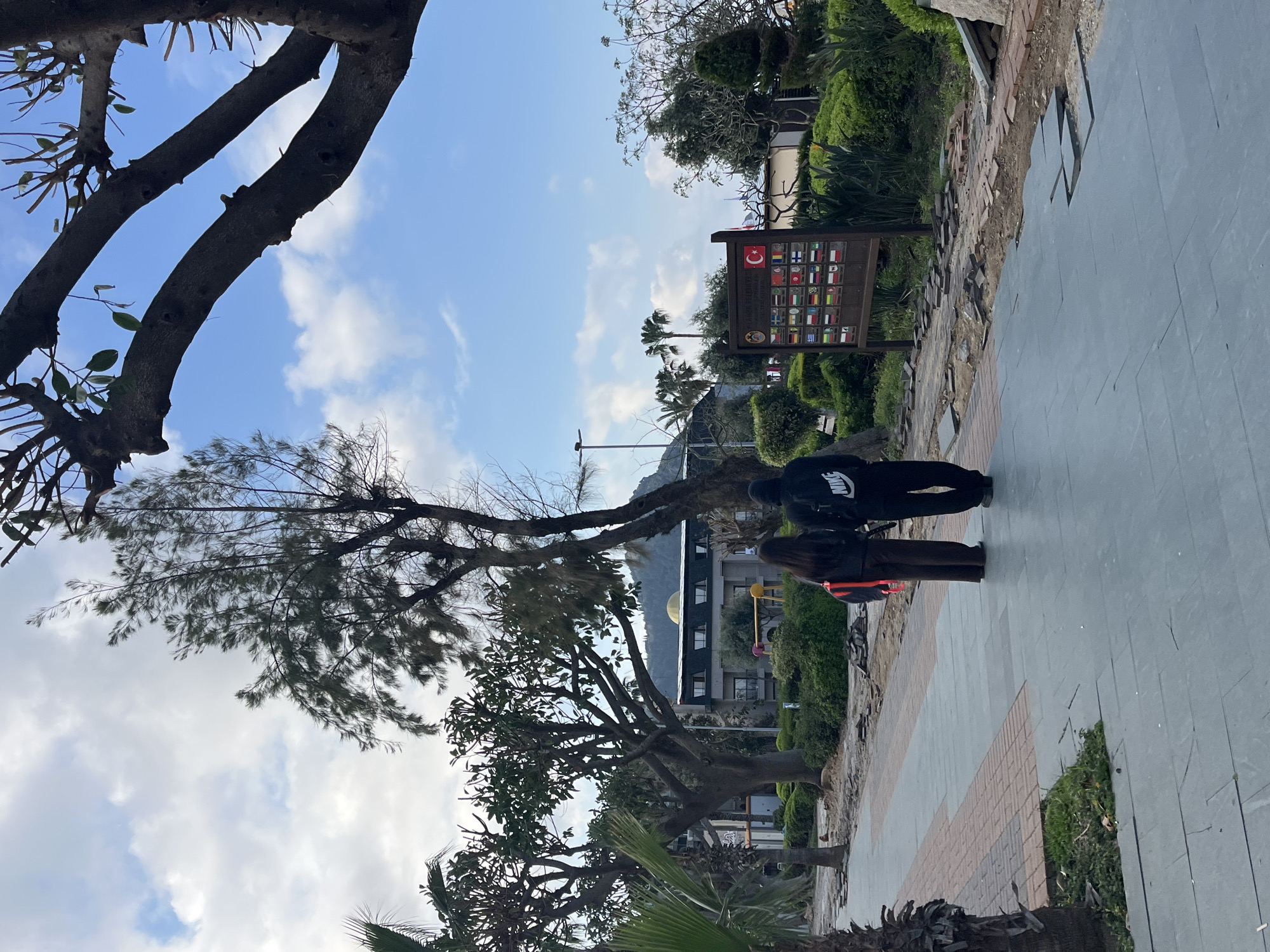 The image captures a park scene in Alanya, Turkey, along a pedestrian walkway paved with grey and brick tiles. Two people, one wearing a "NIKE"-branded hooded jacket, are standing on the walkway. A wooden sign displays the flags of several countries. Green shrubbery and trees surround the walkway, with additional palm trees in the distance. A building with a distinctive golden dome is partially visible. The sky is blue with scattered clouds. The image appears to have been rotated.