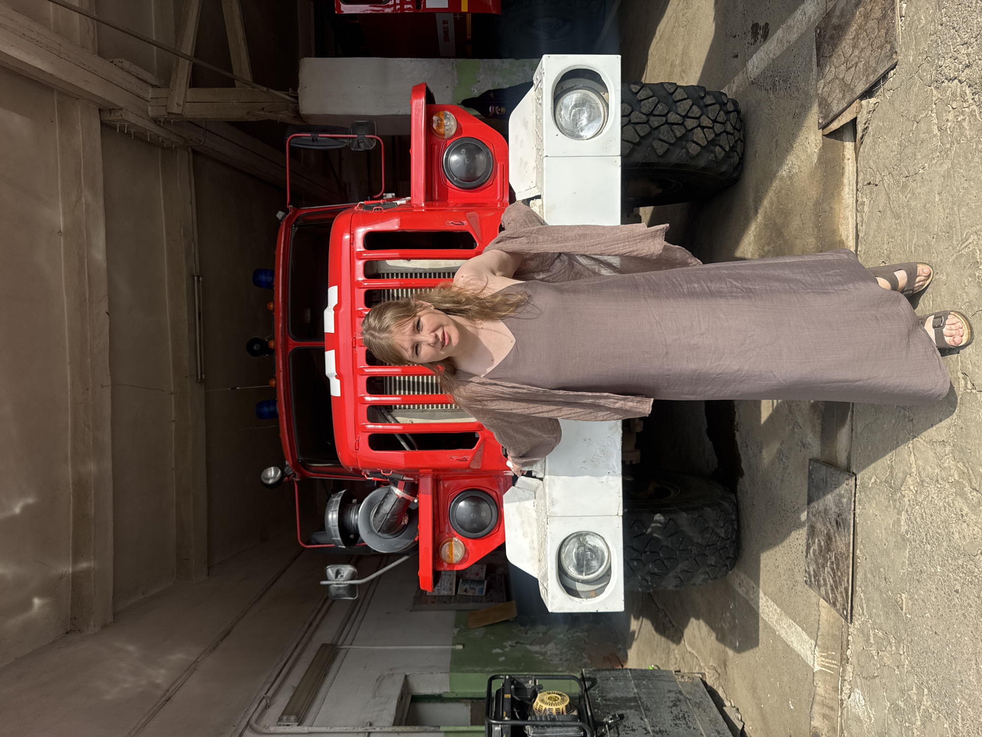 A young woman with blonde hair stands in front of a red fire truck in a garage. She is wearing a long, loose, brown dress and a light brown shawl draped over her shoulders. The fire truck is large and appears to be a heavy-duty vehicle, with large headlights and rugged tires. The garage setting suggests a fire station. The image is taken from a straight-on perspective, with the woman positioned centrally in the frame. The background is dimly lit, with concrete walls and a ceiling visible. The overall impression is one of a posed photograph, likely taken during a visit to the fire station.