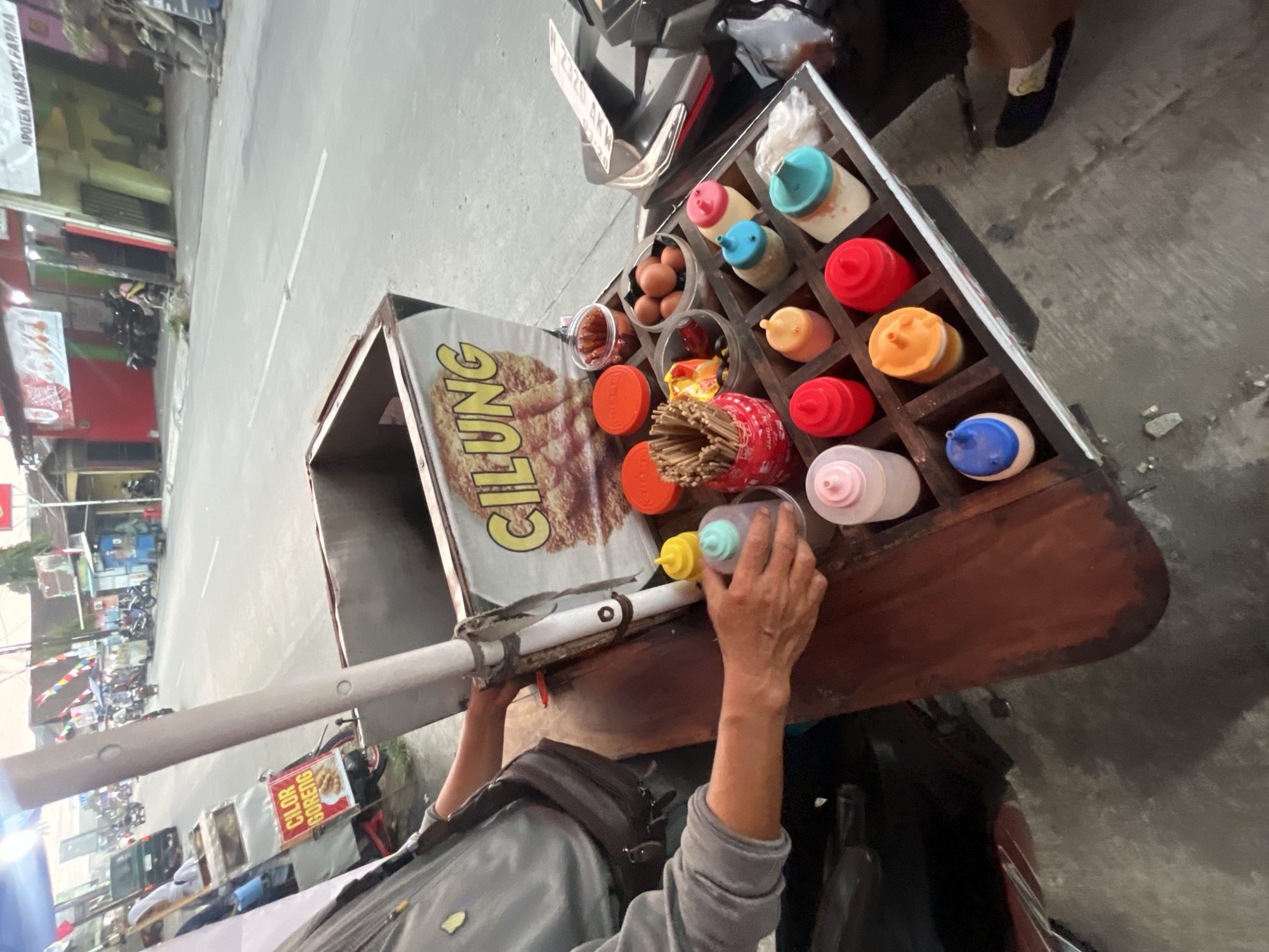 The image captures a street food vendor's cart in Boja, Indonesia. The cart is primarily made of wood and metal, with a prominent sign displaying "CILUNG GORENG." Various condiments are neatly arranged in individual compartments on a wooden tray attached to the cart, including bottles of sauces, eggs, and skewers. A person, presumably the vendor, is seen tending to the cart, with their hands reaching towards a bottle of sauce. In the background, a street scene unfolds with vehicles, shops, and pedestrians, suggesting a busy urban environment. The lighting indicates it is daytime, possibly late afternoon, with some artificial lights also visible.