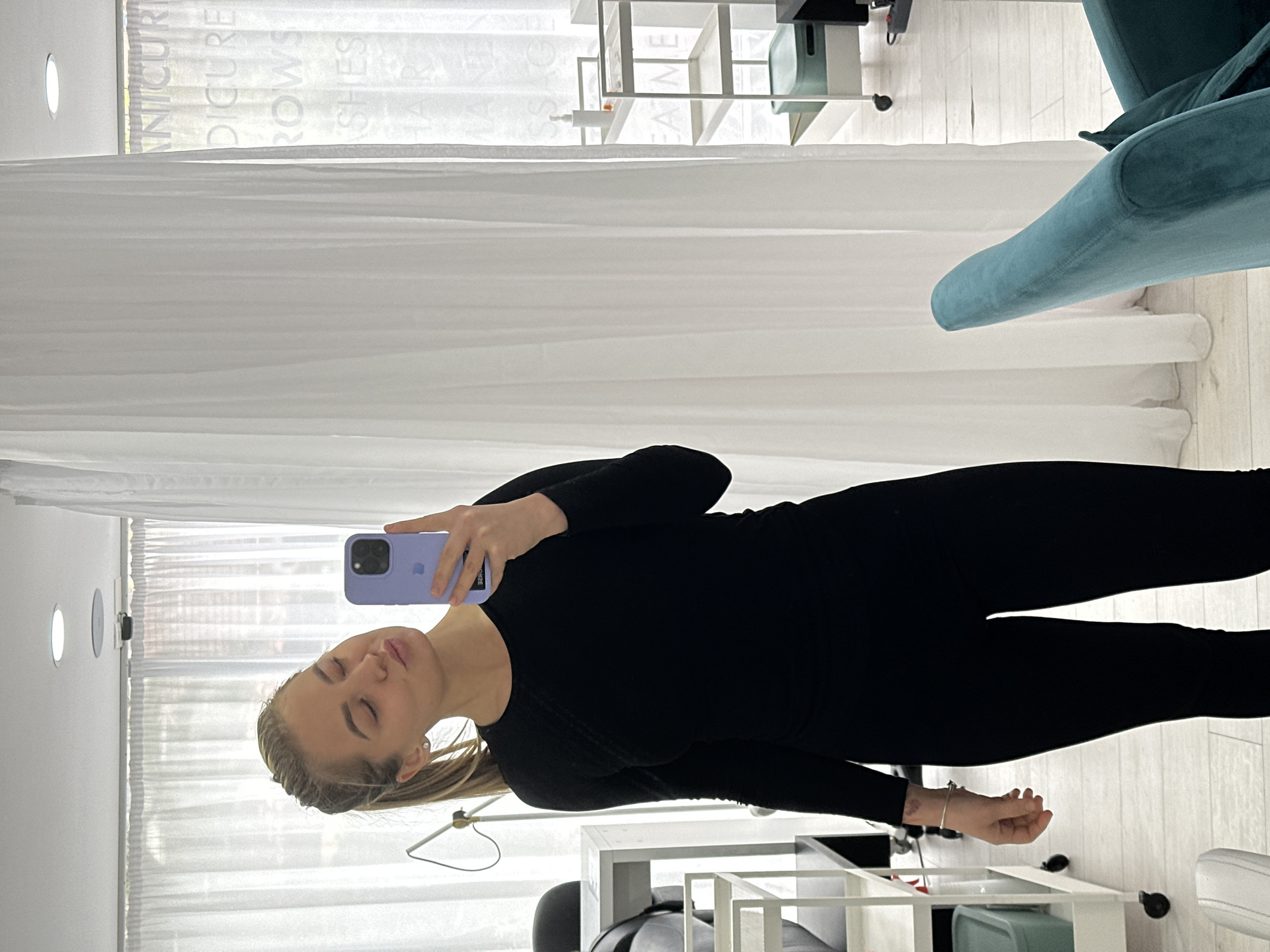A woman in a black outfit is taking a selfie in a well-lit room. The background includes white curtains with printed text and a light blue velvet chair. There are white shelves and treatment tables. The location appears to be a beauty salon or treatment room. There's no clear indication to place the room in Budva, Montenegro.