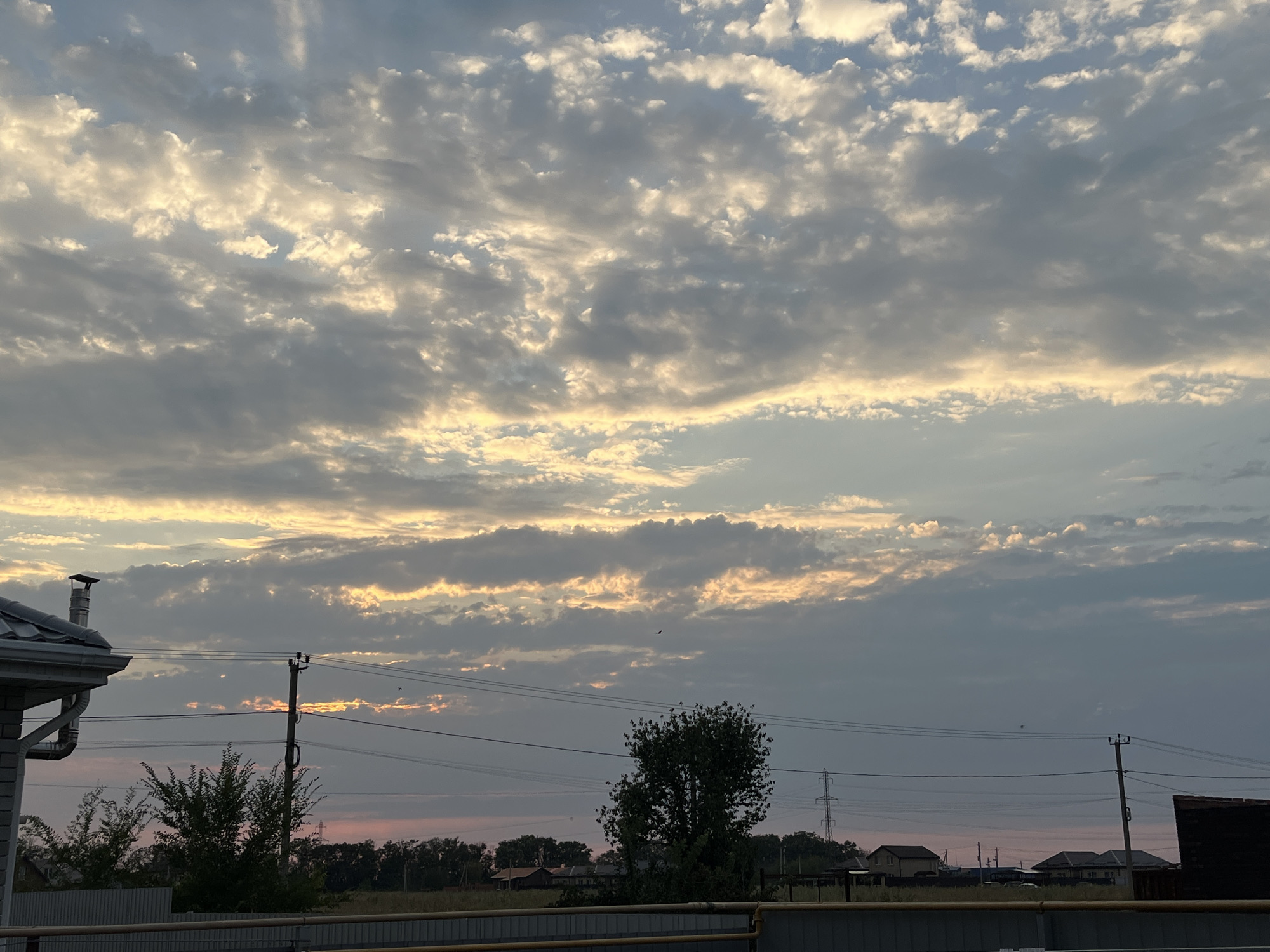 The image depicts a cloudy sky over a rural or suburban landscape. In the foreground, a gray metal fence runs horizontally across the bottom of the frame. Behind the fence, there are trees and houses, with power lines and utility poles visible. The sky is filled with scattered clouds, some of which are illuminated by the setting sun, creating a warm, orange glow. The overall impression is one of a peaceful evening in a quiet residential area. The location is identified as Taganrog City District, Russia.
