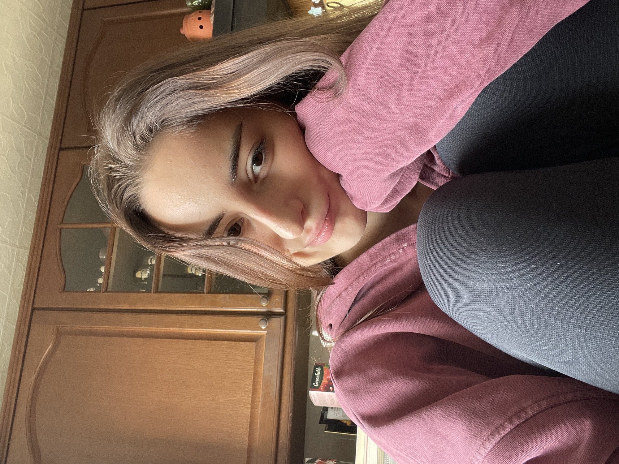 A young woman is positioned in an indoor setting. She is dressed in a pink sweatshirt and dark leggings. Her hair is long and light brown. To the left of the woman is a wooden cabinet.