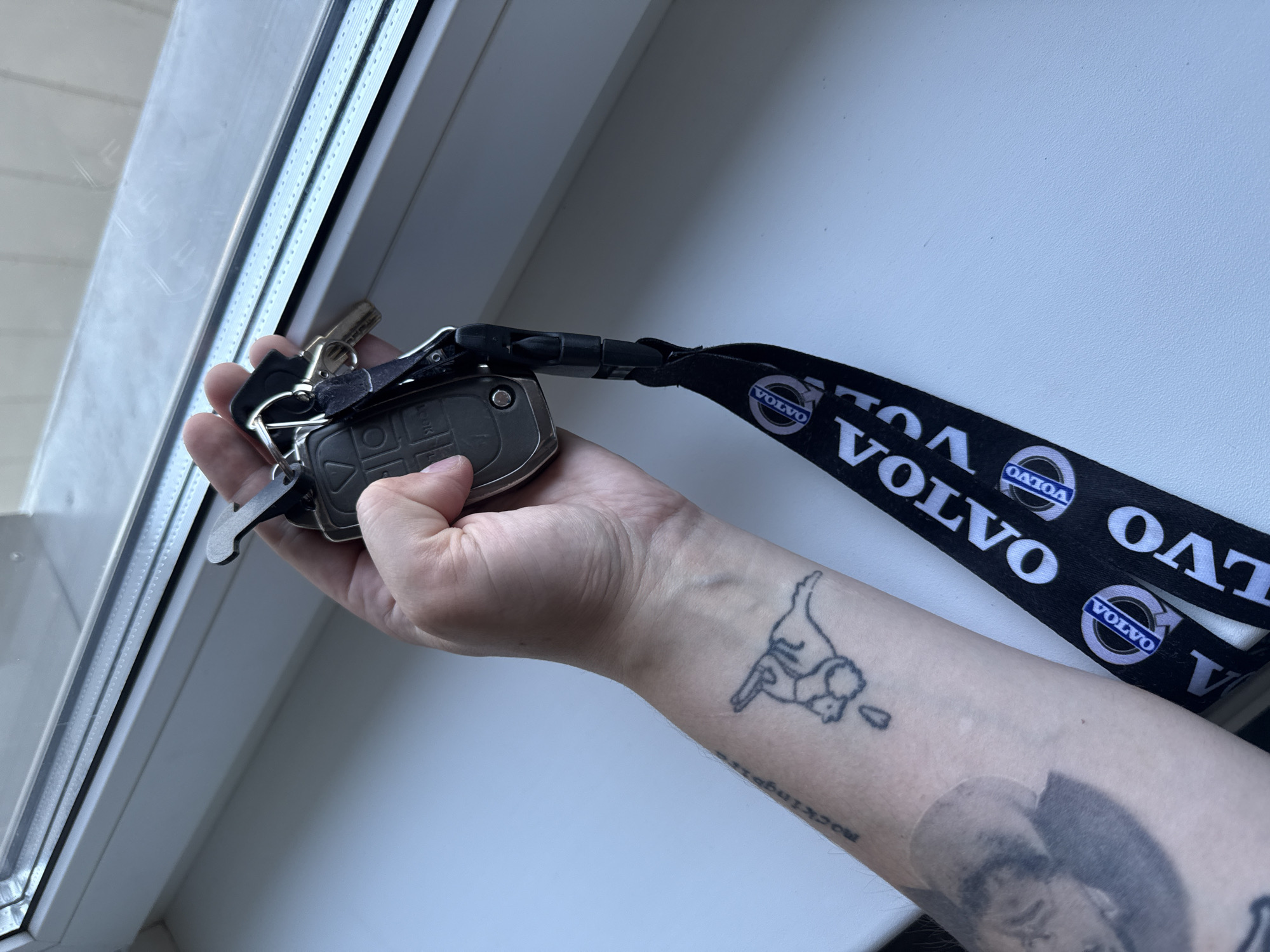 The scene is located in Khimki, Russia. A person's forearm and hand are prominently featured, holding a set of keys over a white window sill. The arm exhibits multiple tattoos: a stylized outline of an animal figure, possibly a rabbit, with a drop below it on the inner forearm; a larger, shaded portrait of a man's head below the animal tattoo; and partially visible text near the wrist, reading "mockingsi". The hand has a light skin tone. The keys include a dark grey Volvo car key fob, multiple metal keys, and a metallic, hook-shaped tool attached to a keyring. A black lanyard, printed with white "VOLVO" text and the Volvo logo, hangs from the key fob down the forearm. The background consists of a bright white window sill and a window with a white frame, reflecting indistinct shapes. Natural light illuminates the scene from the window.