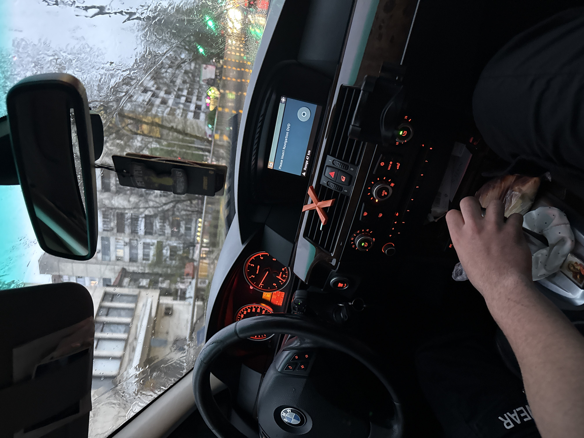 The image captures the interior of a BMW car, viewed from an angled perspective across the driver's seat, looking towards the dashboard and out the windshield. The weather outside is rainy, with numerous water droplets and streaks visible on the windshield, distorting the view of the urban environment. Through the wet glass, several multi-story buildings, characteristic of an urban or suburban area like Burgas, Bulgaria, are visible.
Inside the car, a person's right arm and leg are visible. The hand is positioned near the center console, close to a plastic bag containing what appears to be a snack or food item. The car's dashboard is illuminated with orange lights, including the instrument cluster showing a speedometer and tachometer (both at zero, indicating the car is stationary or off), and the climate control panel with settings like "AUTO" and "MAX". An infotainment screen displays "Please insert Navigation DVD" and "Range 47 km". A small, orange "X"-shaped vent clip is attached to an air vent. Hanging from the rearview mirror is an air freshener with "PEW PEW" visible text. Keys are in the ignition. The steering wheel, bearing the BMW logo, is prominent in the foreground. The person's dark clothing has text "EAR" visible.