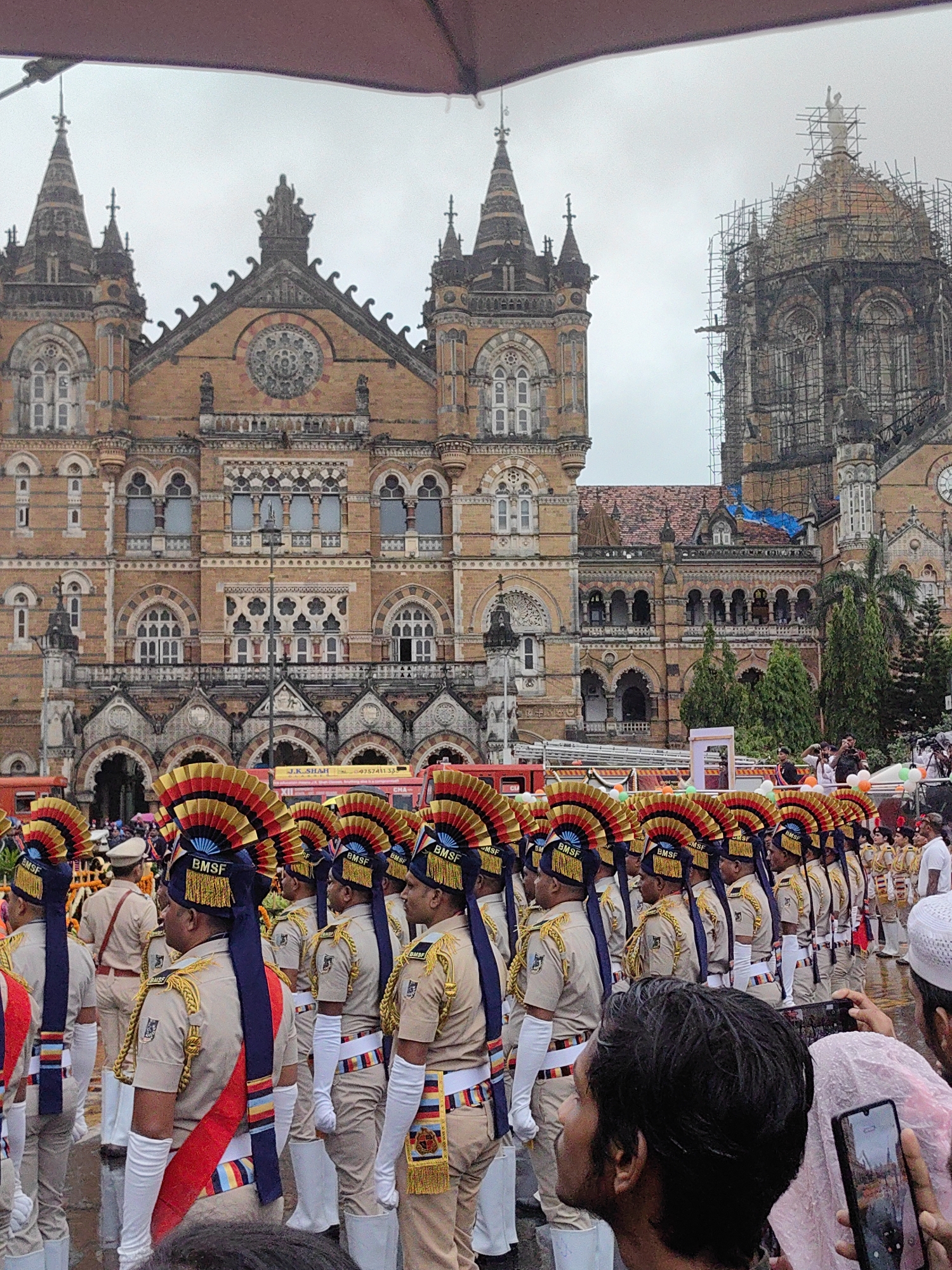 In Mumbai, India, a contingent of uniformed personnel, identified by the "BMSF" insignia on their elaborate headdresses, stands in formation before a grand, ornate building. The building's architecture, characterized by Gothic Revival and Victorian styles, suggests it is a significant historical landmark. The personnel, wearing light-colored uniforms with distinctive red and blue sashes and tall, fan-shaped headdresses adorned with red, yellow, and blue, appear to be participating in a ceremonial event or parade. Their attire and the presence of a large crowd in the background indicate a public gathering. The sky is overcast, and the overall lighting suggests a cloudy day, possibly with rain given the presence of a red umbrella canopy partially visible at the top of the frame. The building's facade displays intricate stonework, arched windows, and decorative elements, with one section on the right appearing to be under scaffolding and renovation. The scene captures a moment of official ceremony against the backdrop of a culturally significant architectural site in Mumbai.