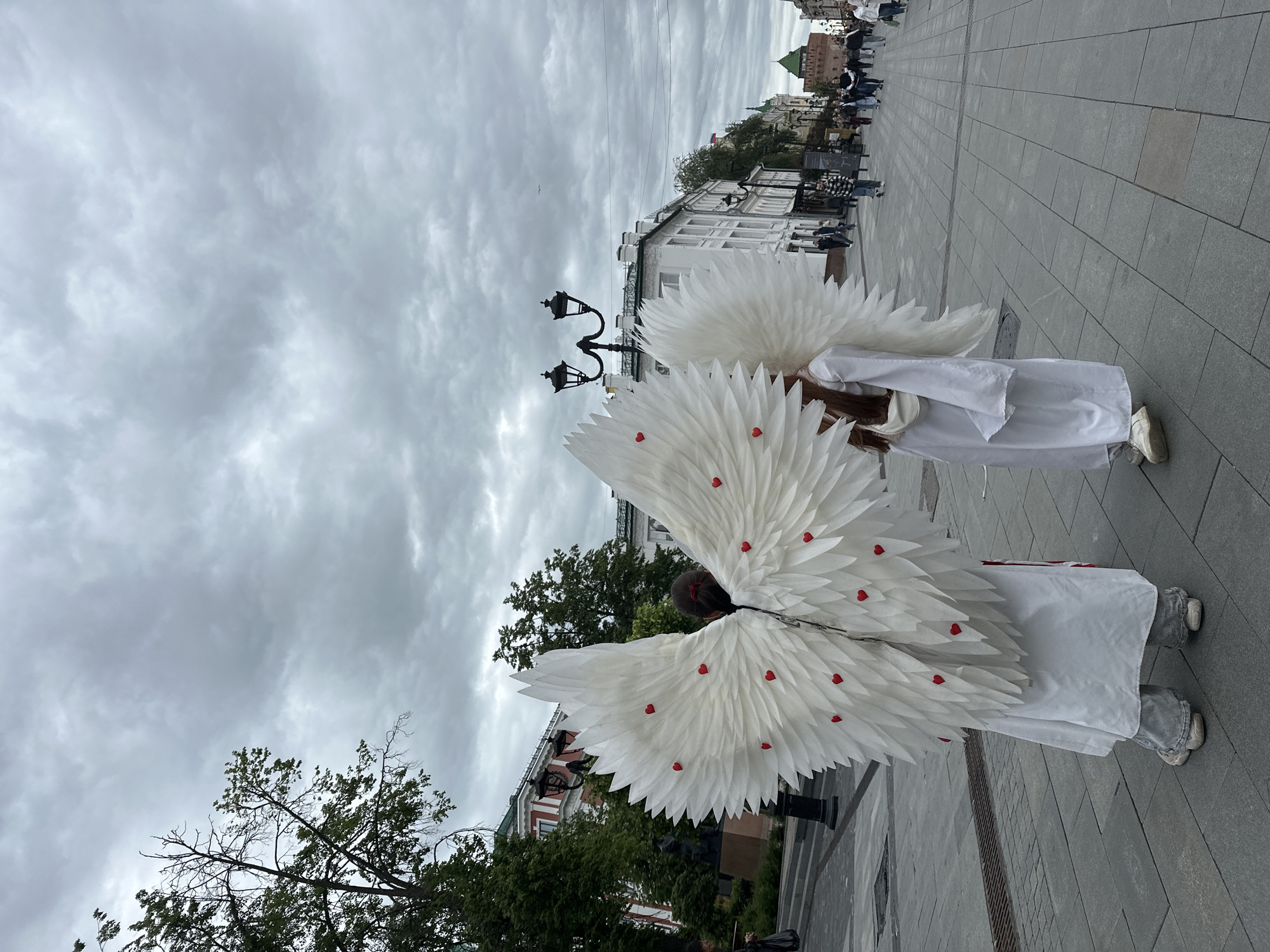 Two individuals are seen from behind, wearing large white wings adorned with small red hearts. They are dressed in white robes and stand on a paved pedestrian walkway. In the background, there are buildings with white facades, trees, a street lamp, and people walking in the distance. The sky is overcast. The location is the pedestrian street Bolshaya Pokrovskaya in Nizhny Novgorod, Russia.