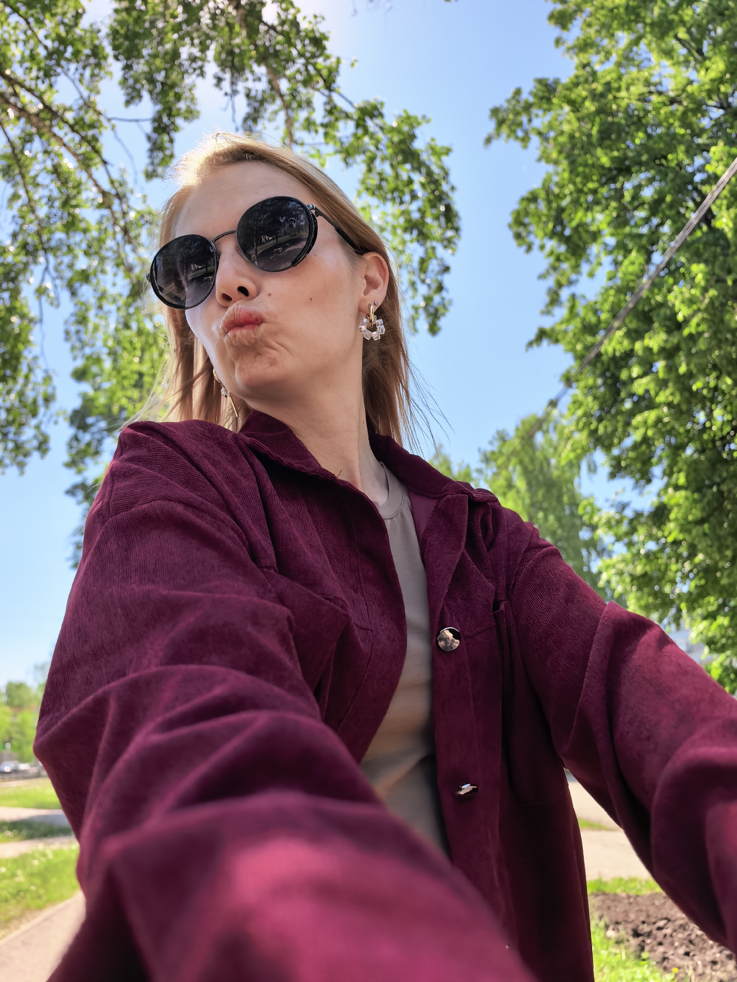 A fair-skinned woman with light blonde hair and wearing round black sunglasses is posing for a selfie outdoors. She is wearing a dark red, corduroy jacket over a light-colored shirt and dangly earrings. She is making a "duck face" expression. The background consists of trees, a blue sky, and a green space that may be a park or lawn. The location is Sterlitamak, Russia. A section of sidewalk and a road are also visible in the background.