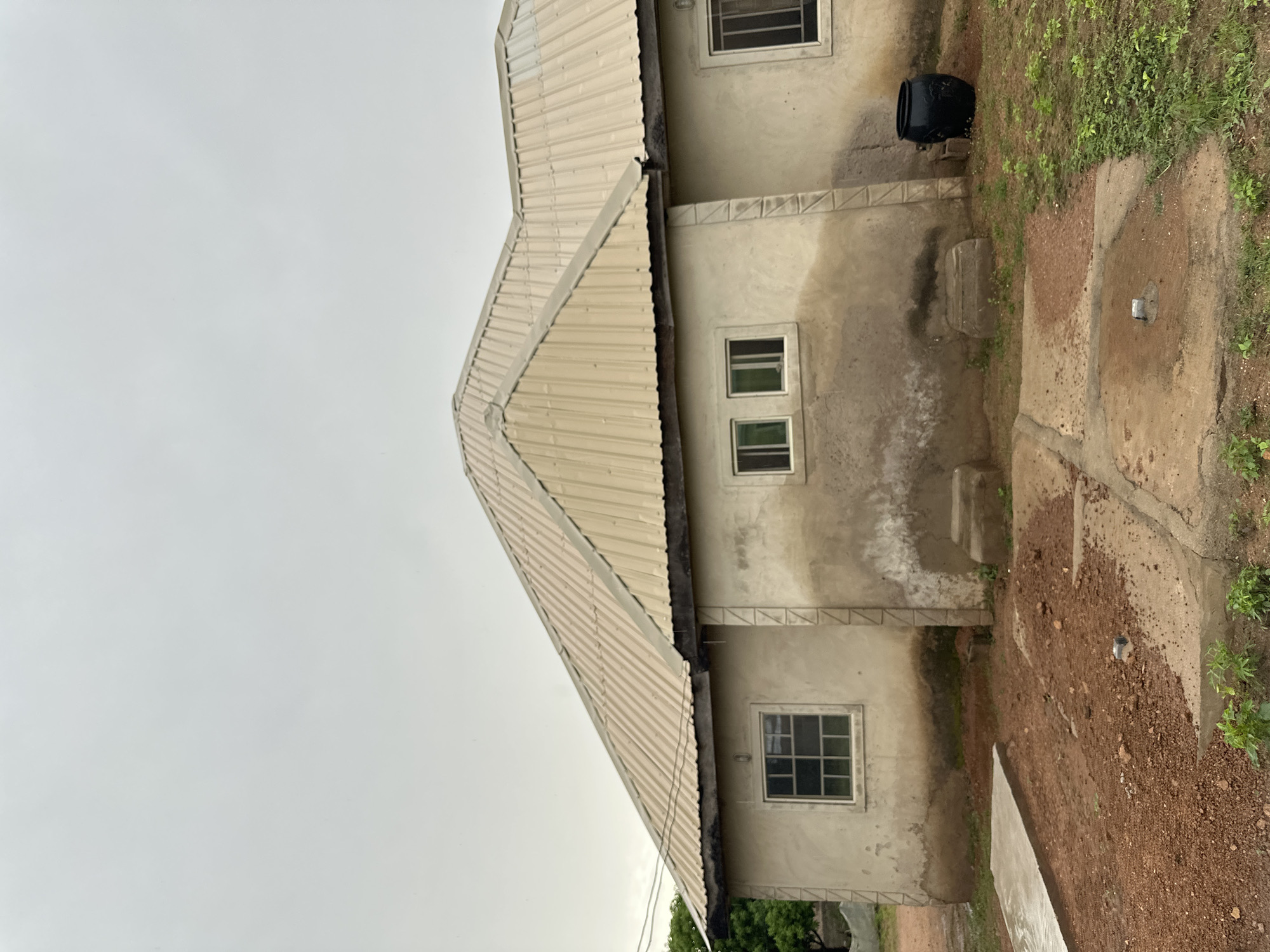 The image shows the exterior of a light-colored, unfinished house in Jalingo, Nigeria. The house has a corrugated metal roof and several windows with metal bars. The walls are made of concrete and appear to be stained and weathered. The ground around the house is covered in red dirt and patches of grass. There is a concrete slab in the foreground, and a large black pot is visible next to the house on the right side of the image. The sky is overcast and a light gray color. There are no people visible in the image.