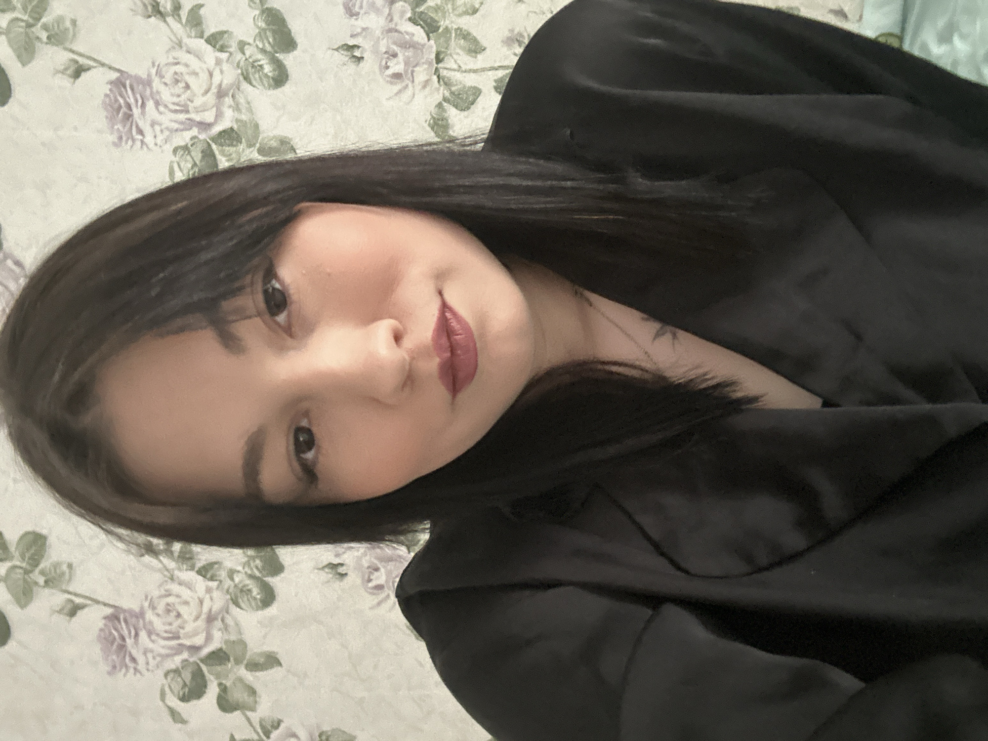The image is a close-up portrait of a woman with dark hair and fair skin. She is wearing a dark, silky garment. Her face is slightly angled towards the camera, and she has a neutral expression. The background is a floral patterned wallpaper with roses. The image does not contain information that would allow for identification of a specific location. The information about Chaikovsky, Russia, is not verifiable from the image.
