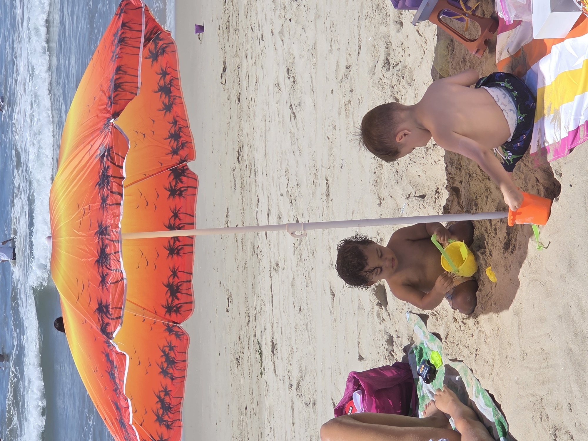 The image depicts a sunny day at a beach, possibly in Santa Monica, USA, with a vibrant orange and red beach umbrella overhead. Two young children are playing in the sand under the umbrella. The child on the left, with curly dark hair, is holding a yellow bucket and appears to be playing with sand. The child on the right, with light brown hair, is wearing patterned shorts and is also engaged in playing with sand, with an orange bucket nearby. A pink beach bag, a green and white striped towel, and various beach toys are scattered around them. In the background, the ocean with gentle waves and a few people swimming can be seen. The sand appears dry and tan, with some areas of darker, damp sand.