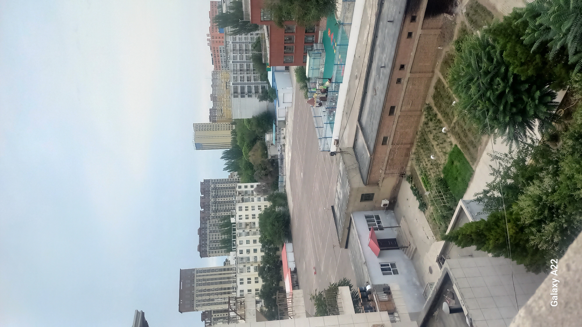 The image is a high-angle view of an urban scene in Korgas City, China, under an overcast sky. In the foreground, a residential building is partially visible, with balconies and windows. Below this, a small garden area with cultivated plots and some greenery is situated next to a brick building. Further into the middle ground, a large, empty paved area, possibly a schoolyard or parking lot, is marked with white lines. Adjacent to this paved area is a children's playground with colorful equipment and a blue fence, as well as some low-lying structures. In the background, a cluster of multi-story apartment buildings and taller residential towers dominate the skyline, interspersed with scattered trees. The overall environment appears to be a mixed-use residential and possibly educational area within a city. There are no people clearly visible in the image.