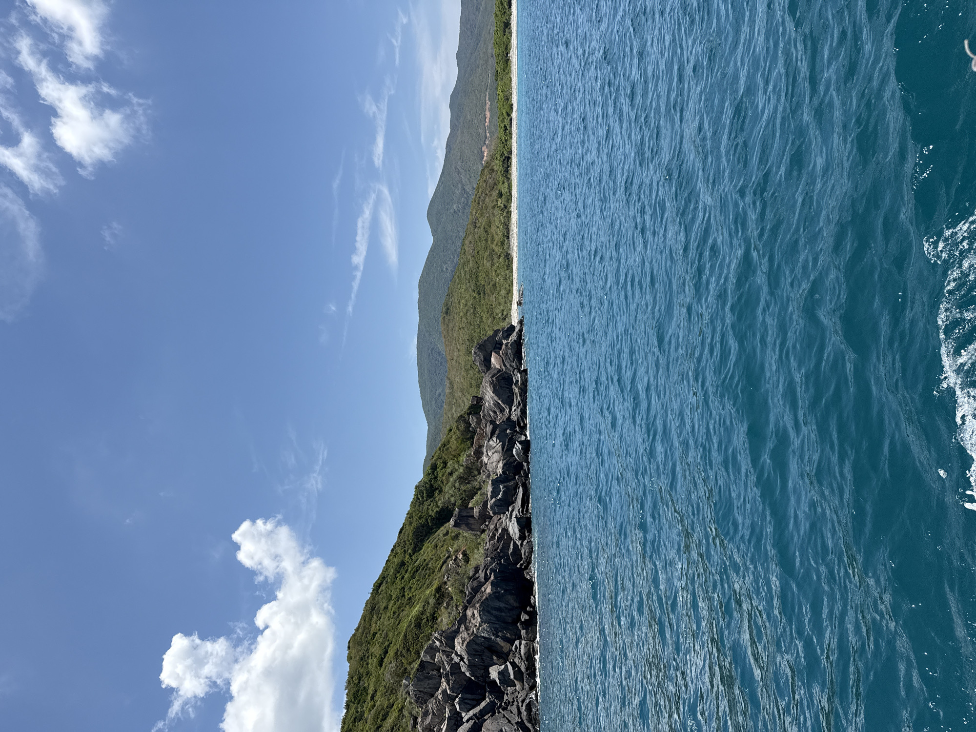 The image shows a coastline with a mountain range covered in green vegetation. The foreground is a rocky shoreline, transitioning to a sandy beach. To the right is the turquoise water of the ocean. The sky is blue with scattered clouds. The location is likely Nha Trang Ward, Vietnam, based on the landscape.