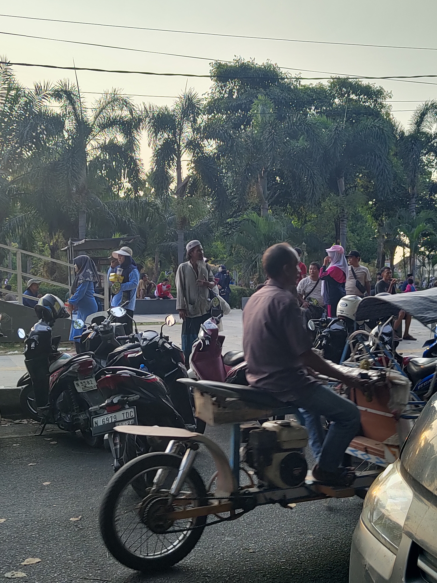 In the city of Bangil, Indonesia, a street scene unfolds with several individuals present. In the foreground, a man is riding a three-wheeled motorized cart. Behind him, multiple motorcycles are parked. Several other people are standing and walking around the area. The background features palm trees and other foliage, suggesting a tropical or subtropical environment. The sky is overcast, indicating either early morning or late afternoon lighting. The overall impression is a typical street scene in a populated area, possibly a market or gathering place.