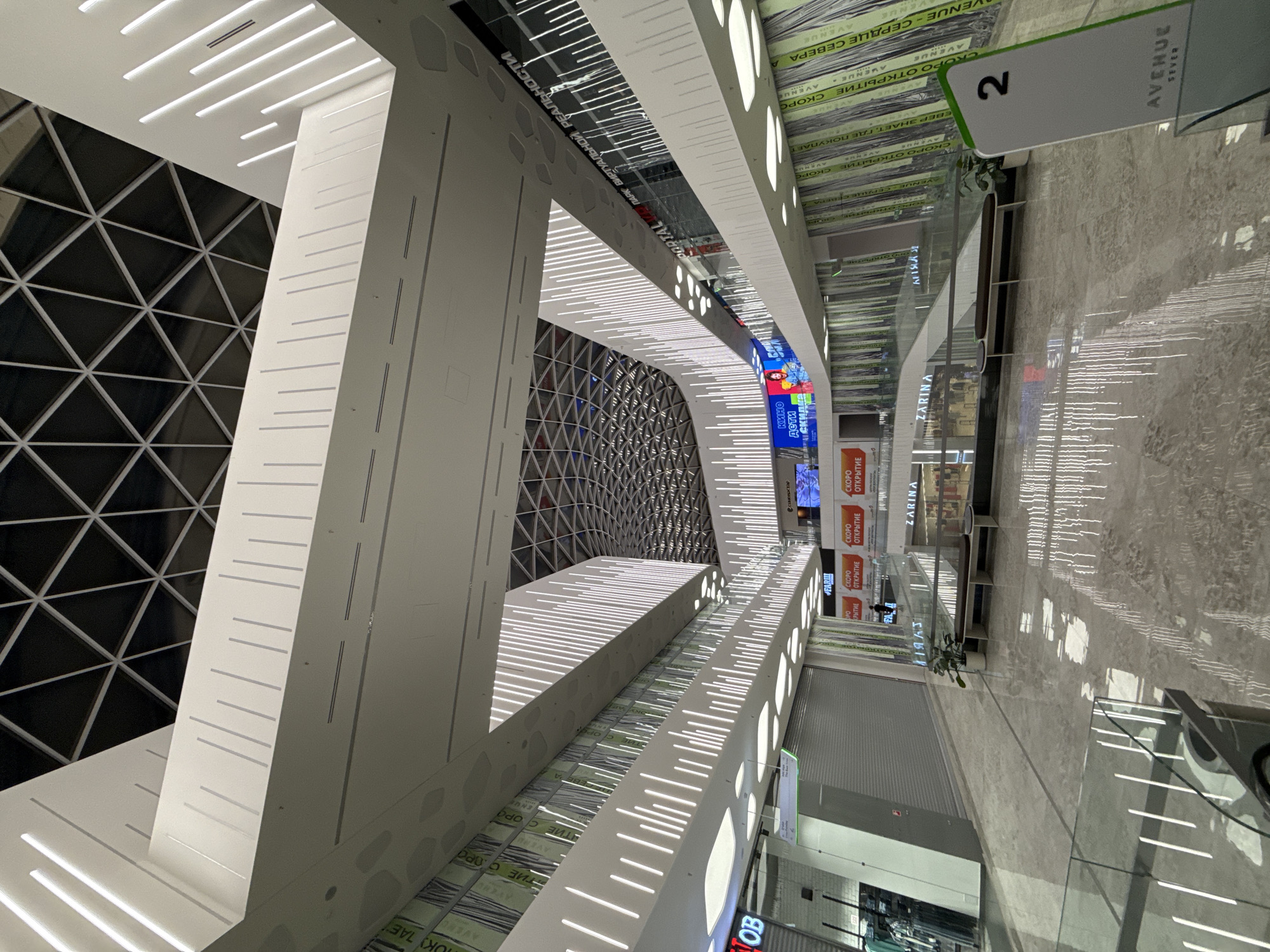 This image presents the interior of the Avenue Sever shopping mall in Moscow, Russia, characterized by modern, multi-level architecture. The perspective is from a lower floor, looking up at the expansive, illuminated space. The ceiling features a prominent dark geometric grid pattern composed of triangular sections. Below this, multiple levels of balconies and walkways are constructed from white, angular forms, extensively decorated with vertical LED light strips that cast reflections on the polished, light-colored floor.
Several storefronts are visible across the various levels. Many display signs reading "СКОРО ОТКРЫТИЕ" (Coming Soon), indicating new businesses or upcoming openings. Specific visible establishments include "ZARINA," a "ПАРК ВИРТУАЛЬНОЙ РЕАЛЬНОСТИ" (Virtual Reality Park), and a "КИНО ДЕТИ" (Kids Cinema). The walls on some levels feature a green and white striped pattern incorporating text, likely branding for "AVENUE SEVER" and "СЕРДЦЕ СЕВЕРА" (Heart of the North). Benches with dark seating and light frames are positioned on the lower level, accompanied by potted green plants. Glass railings line the walkways. A solitary, indistinct individual is visible walking in the mid-ground near a store with a rolled-down shutter. A prominent sign in the foreground displays the number "2" and the text "AVENUE SEVER". Large digital screens on the upper levels show colorful advertisements or media.