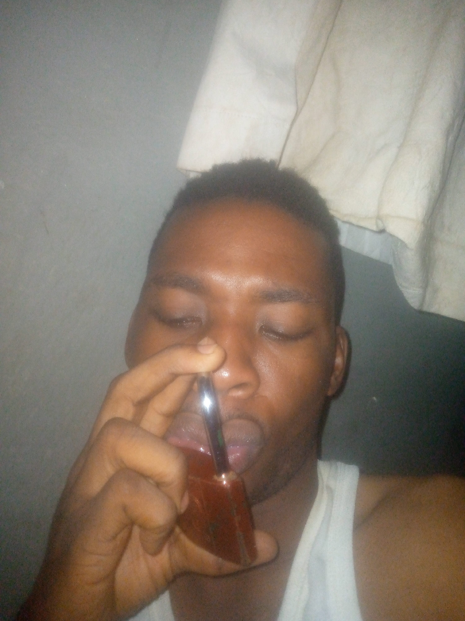 A person is holding a brown glass bottle to their mouth, with a silver straw visible inside the bottle. The person is looking down with their eyes closed. They are wearing a white tank top. The background appears to be a grey wall with a white cloth hanging. The location and city are not identifiable.