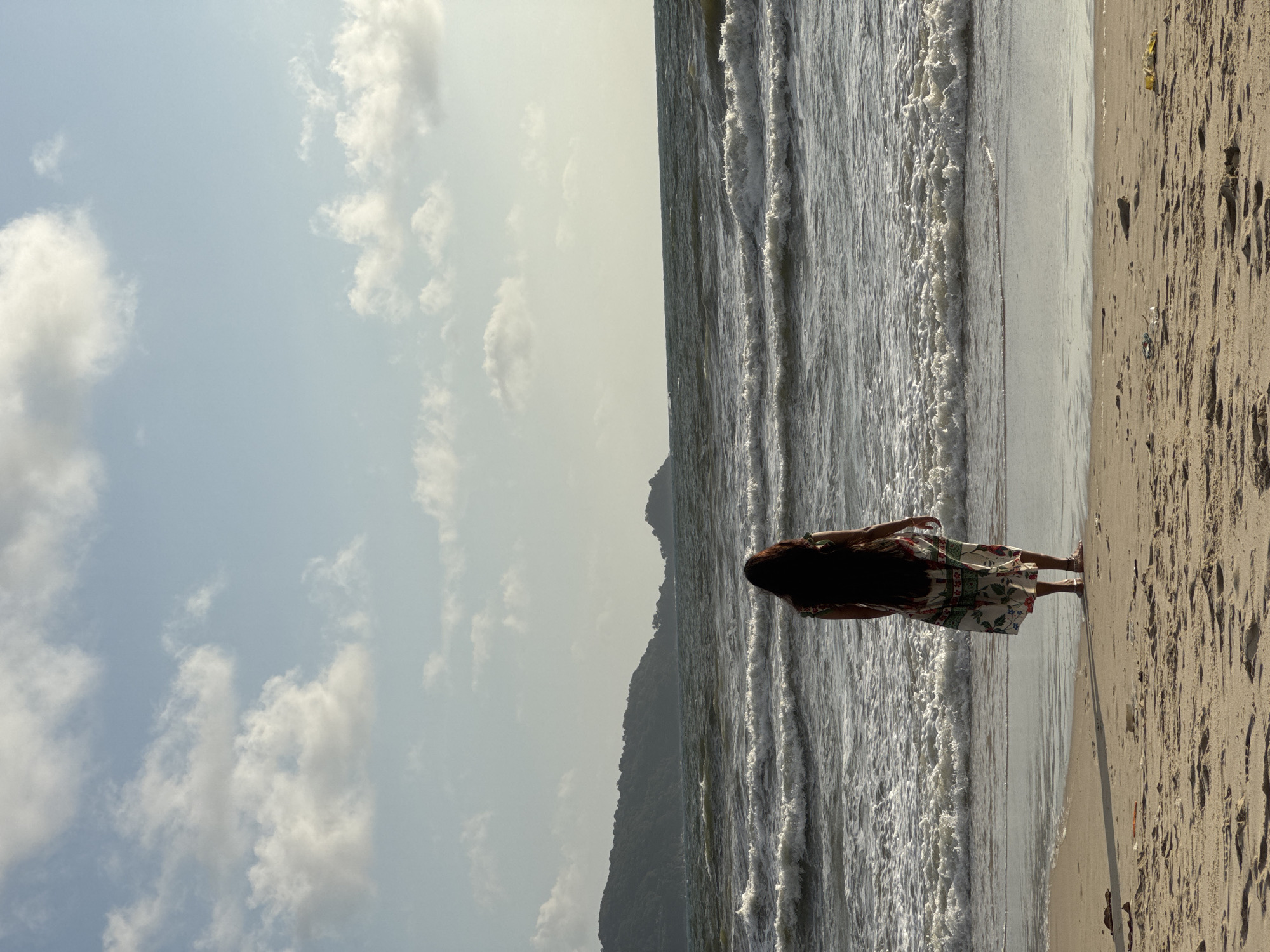 In Patong, Thailand, a lone person stands on a sandy beach facing the ocean. The individual, seen from the back, has long dark hair and is wearing a floral-patterned dress. The waves are moderate, with white foam cresting as they approach the shore. In the distance, a lush, green, mountainous coastline stretches across the horizon under a sky filled with scattered clouds. The sunlight casts shadows on the wet sand at the water's edge, and a gentle breeze appears to be blowing. The overall scene evokes a sense of tranquility and natural beauty.