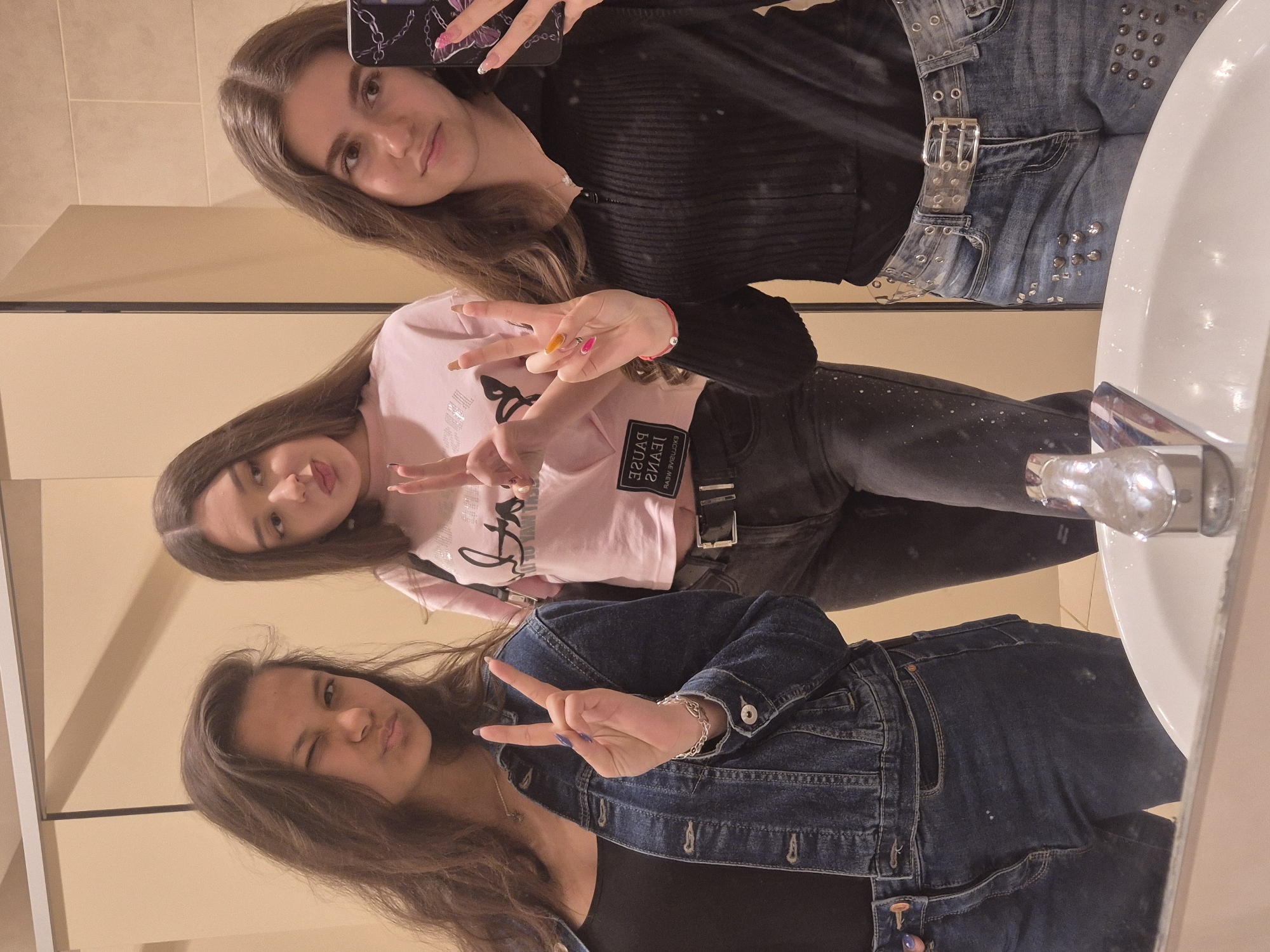 The original media file is a mirror selfie taken by three young women in what appears to be a bathroom setting, specifically in Zagore, Bulgaria. The image is rotated 90 degrees counter-clockwise.
When viewed upright, the scene features three young women posing for the camera.
The woman on the left has long, straight, dark brown hair and looks directly at the camera with a soft smile. She wears a black ribbed long-sleeved top and light blue jeans featuring silver studs and a distinctive clear belt with a silver buckle. She is holding a smartphone with a black case adorned with a silver chain pattern, which is being used to capture the image. Her nails are painted a light pink.
The middle woman has long, wavy, dark brown hair and is making a peace sign with her right hand, showcasing multi-colored nails (orange, pink, and brown/nude). She is wearing a light pink short-sleeved top on which mirrored text "PAUSE JEANS" is visible. She also wears dark-colored pants and a black belt. A thin red bracelet with a silver charm is on her wrist, and she looks towards the camera with a slight pout and gentle smile.
The woman on the right has long, dark brown hair and strikes a playful pose, winking and pouting while flashing a peace sign with her left hand. Her nails are painted a dark blue or purple. She is dressed in a black top underneath a blue denim jacket and dark blue jeans. A silver chain bracelet is visible on her left wrist.
The setting appears to be a bathroom, indicated by a white sink visible on the right side of the frame and light-colored tiled walls in the background. The overall mood is casual and friendly, with the subjects displaying expressions of enjoyment and playfulness.