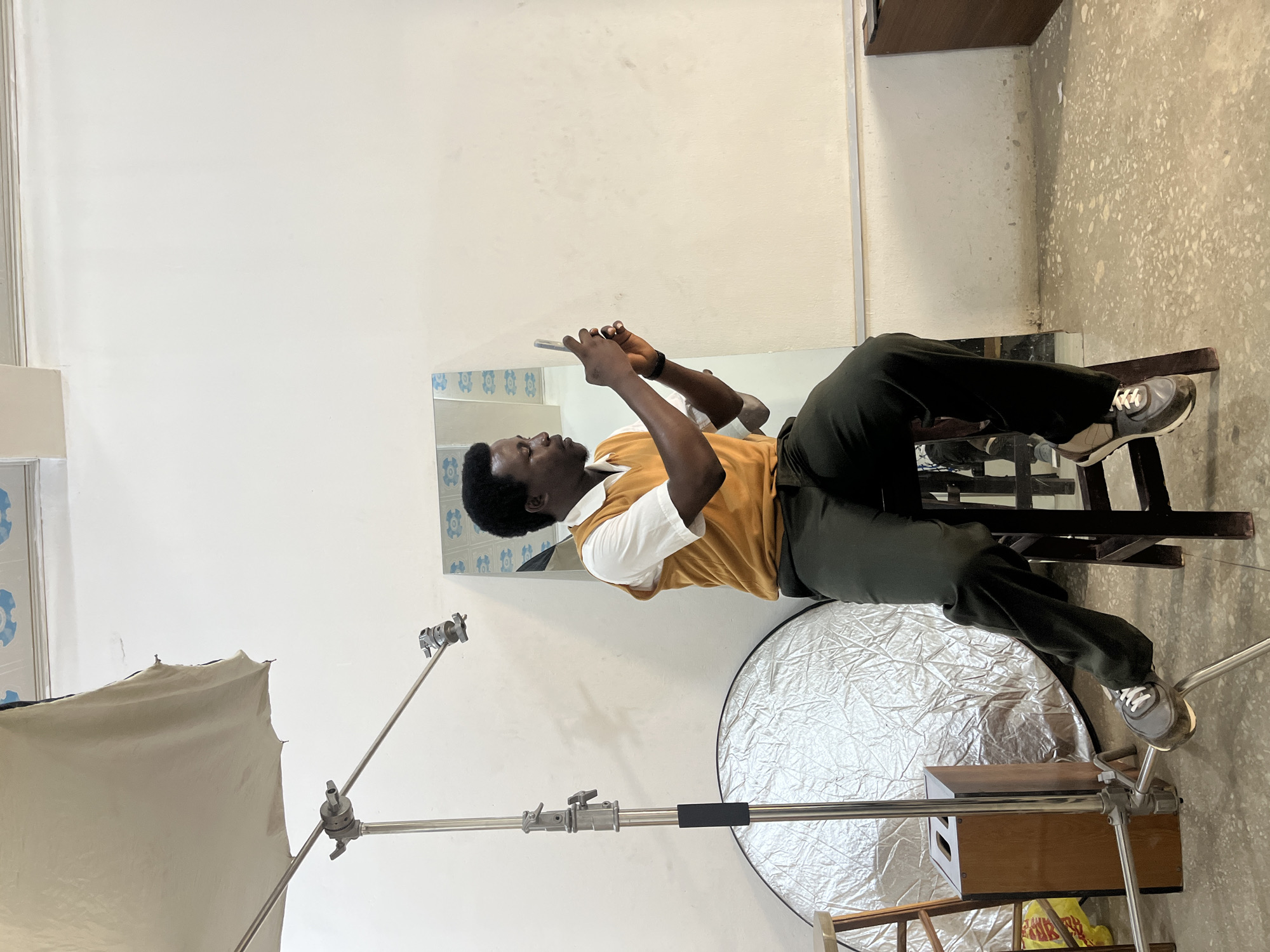 A young Black man is lying on his back on a chair, looking at his phone. He is wearing a yellow shirt with a white collar and dark green pants. He has on sneakers and a black watch. The room appears to be a studio, with a white wall, a large reflector on the floor, and a lighting stand with a diffusion umbrella. A mirror is visible on the wall behind the man, reflecting more of the room and a pattern of blue tiles. The scene suggests a break or downtime during a photoshoot or filming session. The lighting is bright, indicating it's daytime, and there are no obvious weather cues. The overall mood seems relaxed, with the man engrossed in his phone. No visible text is present in the image.