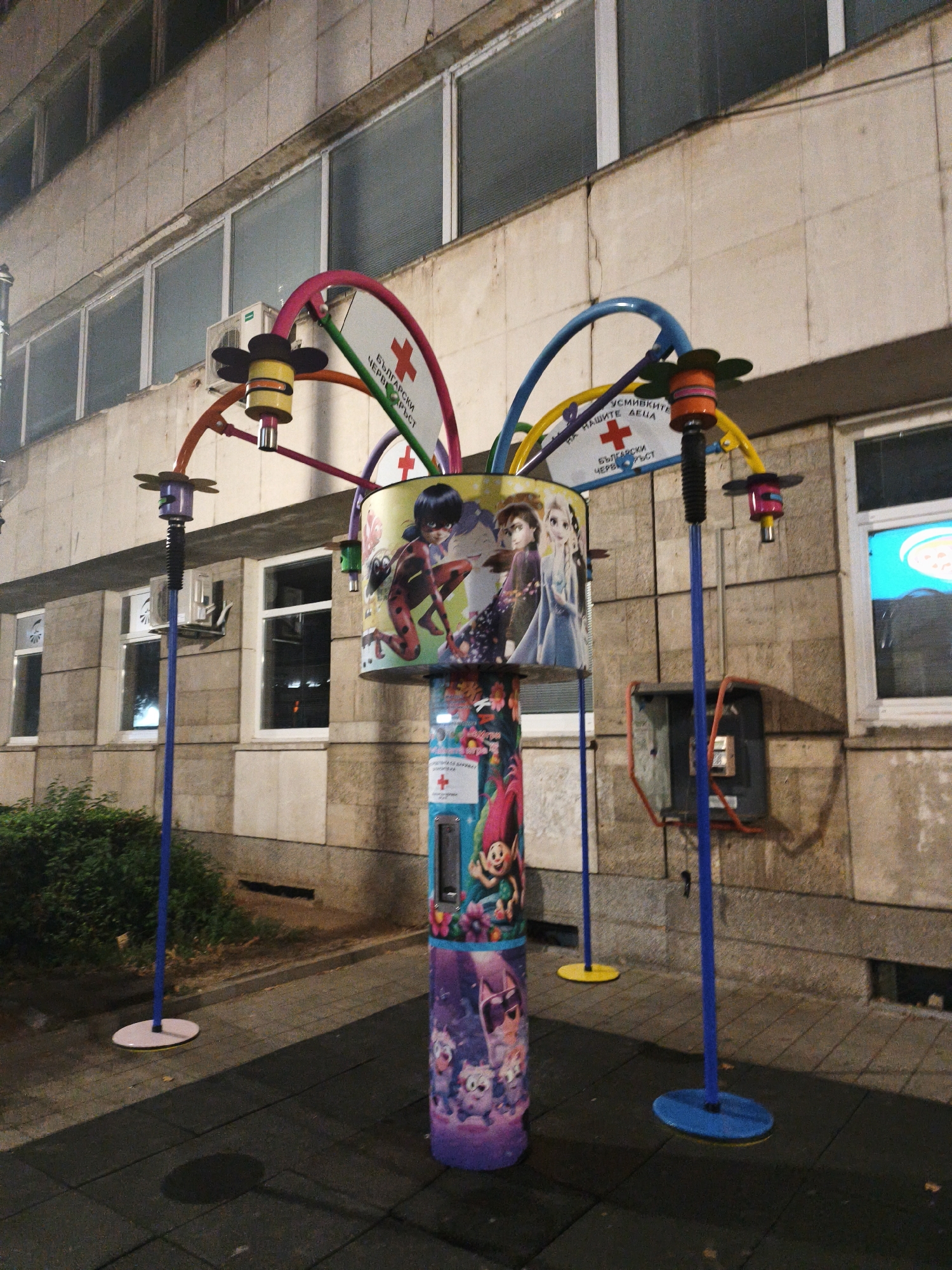 The image shows an outdoor art installation in front of a building in Lovech, Bulgaria. The installation is a colorful, multi-tiered structure with various cartoon characters depicted on its surfaces, including characters from "Miraculous Ladybug" and "Frozen," as well as trolls. The structure is supported by blue poles, and adorned with colorful arches and lights. Two white signs with red crosses are visible at the top of the installation, possibly indicating a connection to a medical or charitable cause. The building behind the installation appears to be an office building or clinic, with rows of windows and air conditioning units. The ground is paved with dark tiles, and a small patch of greenery is visible on the left side of the image. A vintage red and gray telephone booth is mounted on the wall to the right of the installation. The lighting suggests it is nighttime or dusk.