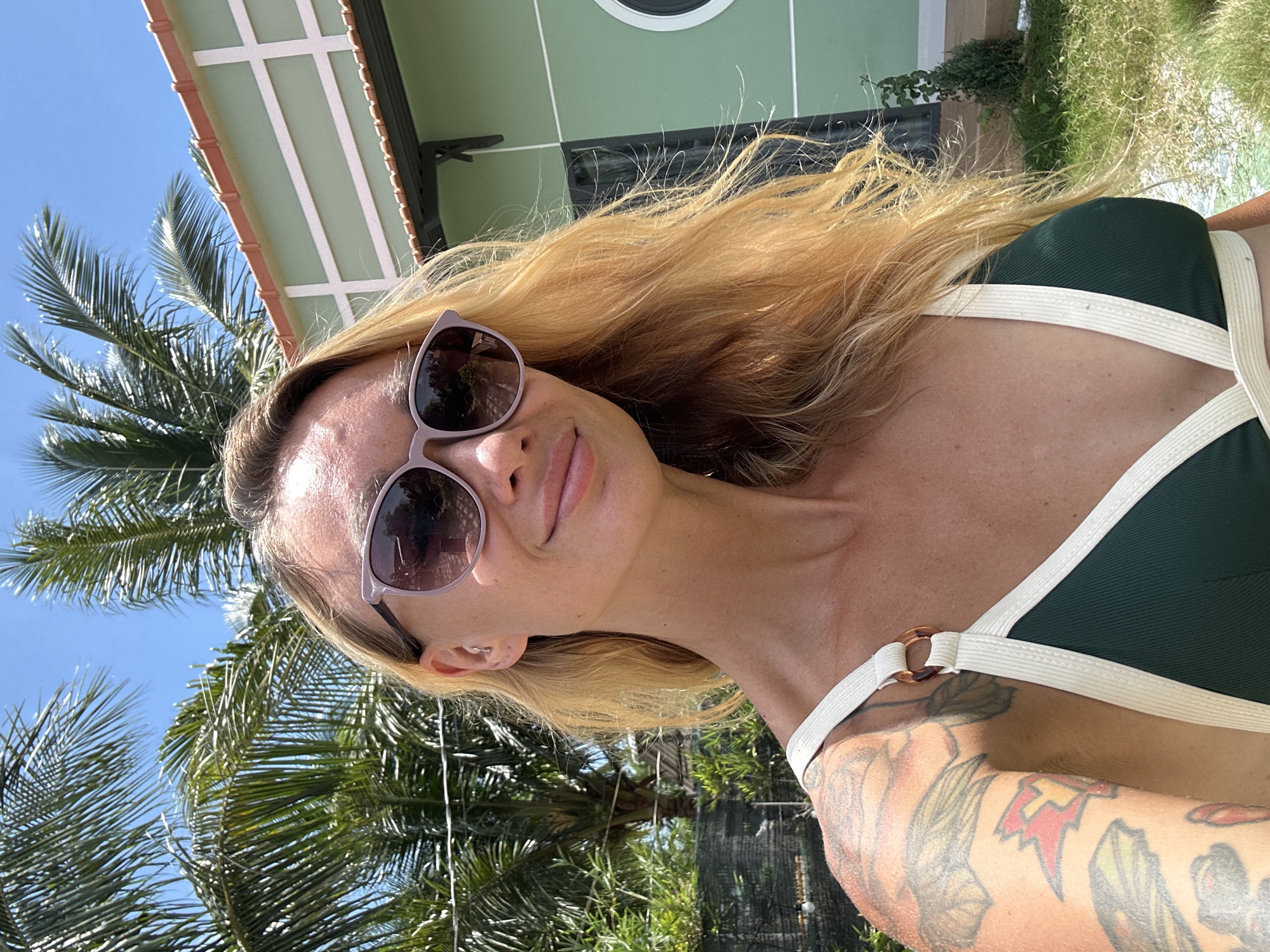 A woman with blonde hair and tattoos on her arm is taking a selfie outdoors. She is wearing sunglasses and a dark green bikini with white trim. Behind her, palm trees sway under a blue sky, and a light green building with white trim is visible. The scene appears to be in Mũi Né, Vietnam.