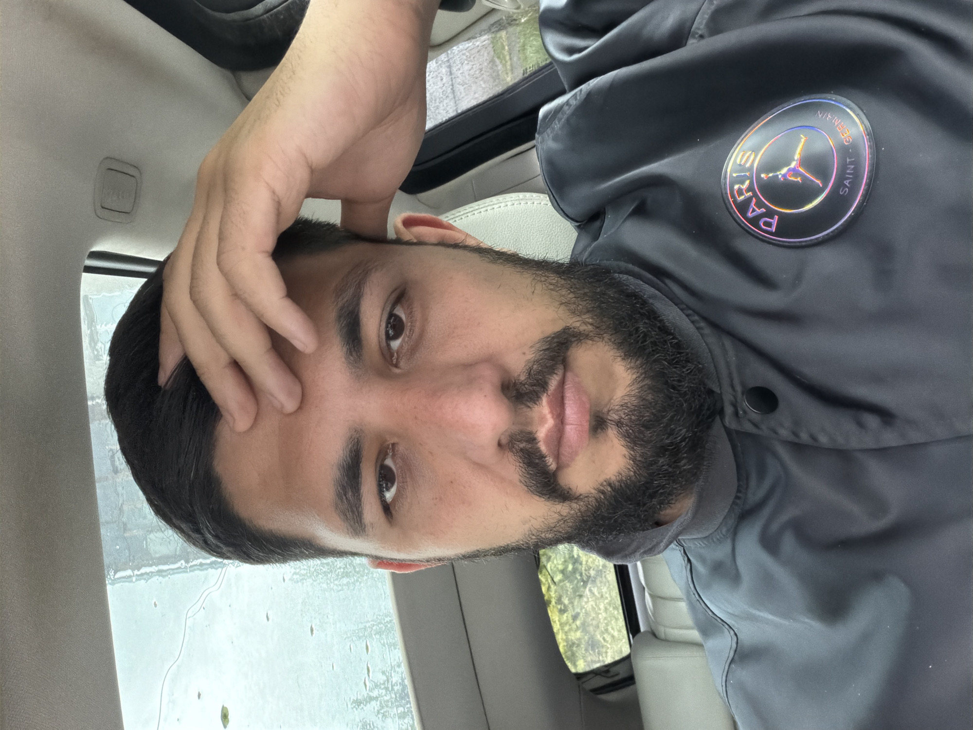 A person with a beard is sitting in a car. The person is wearing a grey jacket with a Paris Saint-Germain logo. It appears to be raining outside. The location is indeterminable.