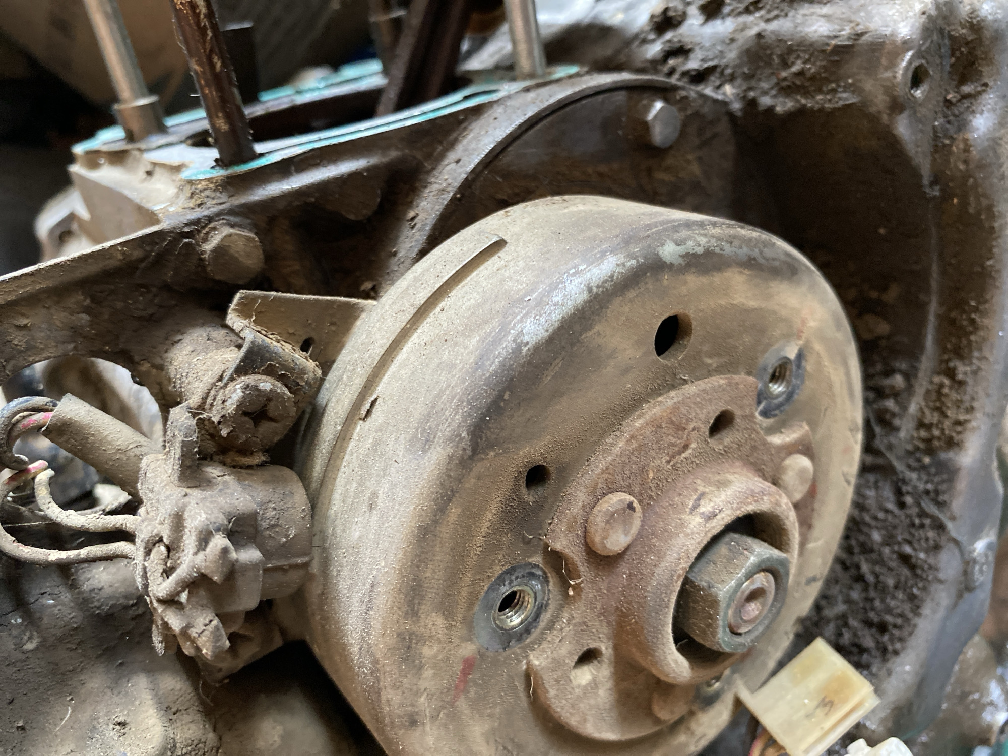 The image is a close-up, slightly angled shot of a heavily soiled engine component, possibly from a motorcycle or small engine. The primary subject is a rusty, circular metal piece with a large central nut and several smaller bolts or screw heads around its perimeter. The surface is covered in thick dust and grime. To the left, a cluster of wires is visible, connected to a weathered electrical connector that is also coated in dirt. In the background, other engine parts are visible, along with some metallic rods and possibly some debris. The overall environment appears to be a workshop or storage area, given the presence of mechanical parts and the accumulation of dirt and dust. There are no people or animals present in the image. The lighting is somewhat dim, and the focus is on the details of the engine component.