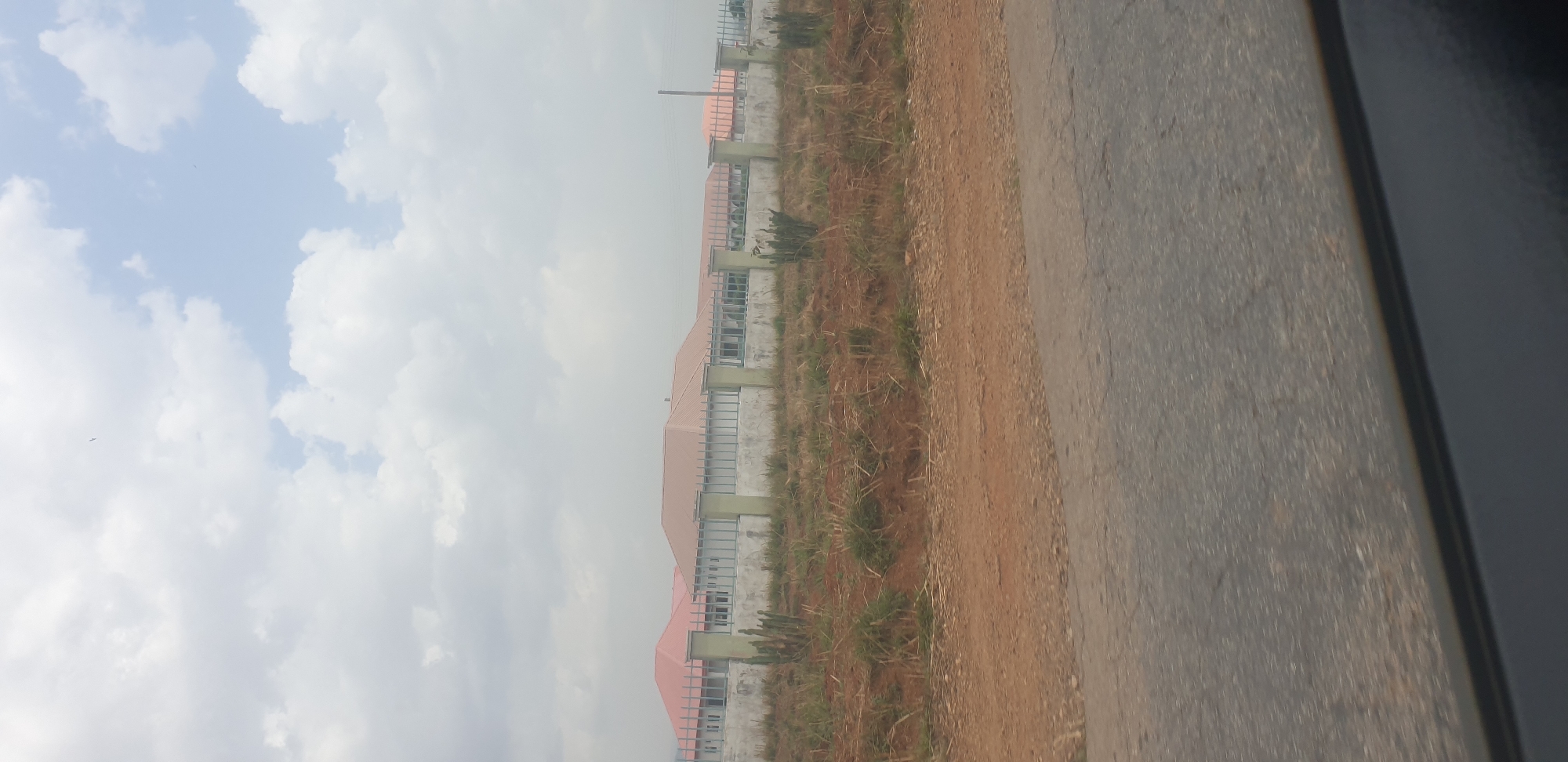 The image presents an outdoor scene in what appears to be Hoss, Nigeria, under a partly cloudy sky. The perspective is from ground level, likely from or near a road.
In the foreground, on the left side of the frame, there is a dark grey, paved surface, resembling an asphalt road. Adjacent to this road is a strip of reddish-brown, unpaved earth, sparsely covered with dry, light-colored grass and some small green shrubs.
Further back, a row of multiple buildings stretches across the mid-ground. These structures have uniform designs with distinctive reddish-brown, possibly corrugated metal, roofs. The walls appear to be made of light-colored concrete or block, featuring numerous window openings, some fitted with horizontal grates or frames. The buildings suggest a planned development or residential/commercial area.
The upper portion of the image is dominated by the sky, filled with scattered white and light grey cumulus clouds, with patches of lighter blue visible between them. The overall lighting indicates it is daytime, possibly an overcast or partly cloudy day.
No people or animals are visible, and there is no specific activity taking place. The scene is still and observational. The rightmost edge of the original image shows a dark, indistinct area, possibly a part of a car's interior or window frame from where the photo was taken. No visible text is present in the image.
