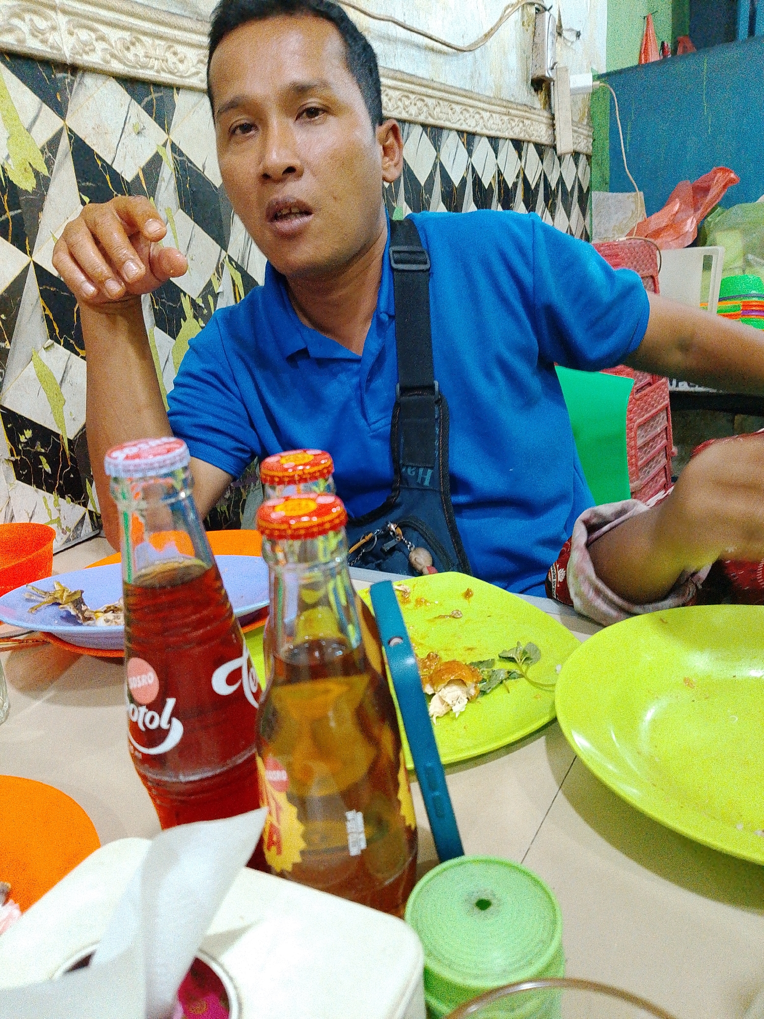 A man in a blue polo shirt is seated at a table in an outdoor eating establishment in Bandar Setia, Indonesia. He is looking directly at the camera with an open mouth, and his left hand is raised with his index finger extended. In front of him are two glass bottles of soda, one red and one amber, with red and yellow caps. There are also two green plates on the table, one with remnants of food and a small sprig of greenery, and a white tissue box with a tissue partially pulled out. The table is covered with a light-colored surface, and the background wall is decorated with a geometric pattern of black, white, and green tiles. A strap from a bag is visible across the man's chest. The overall setting appears to be casual and unpretentious.