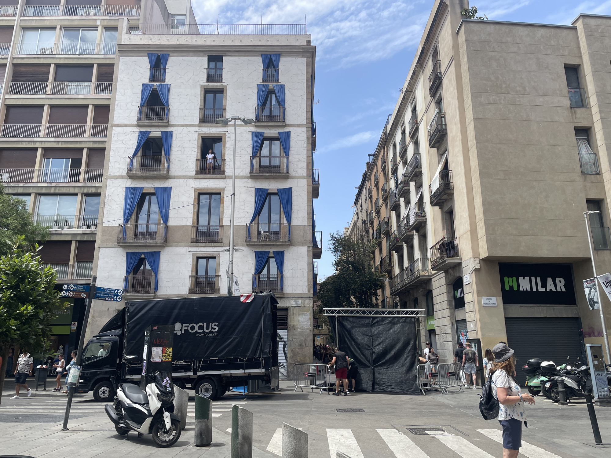 This is a medium-shot, eye-level photograph taken on a sunny day in Barcelona, Spain. The scene depicts a street corner with multiple buildings and a street. In the foreground, a white scooter is parked on the sidewalk. A black truck with the logo "FOCUS" and website "www.focus.cat" is parked on the street, partially blocking a crosswalk.
The buildings in the background feature balconies with blue curtains visible on the facade of the building on the left. A few people are visible on the street, some walking, and others standing near the truck and a black tent structure. One person is on a balcony of the building on the left, looking out. The overall atmosphere seems casual and bustling, with a mix of residents and workers.
Visible text includes "MANA", "HOLA", "CAP", "VEINA", "FORA DEL", "BARRI!", "FOCUS", "www.focus.cat", "FRANCESC CAND", and "MILAR". The presence of a truck and tent suggests a possible event or setup in progress. The sky is partly cloudy, indicating a pleasant daytime weather.