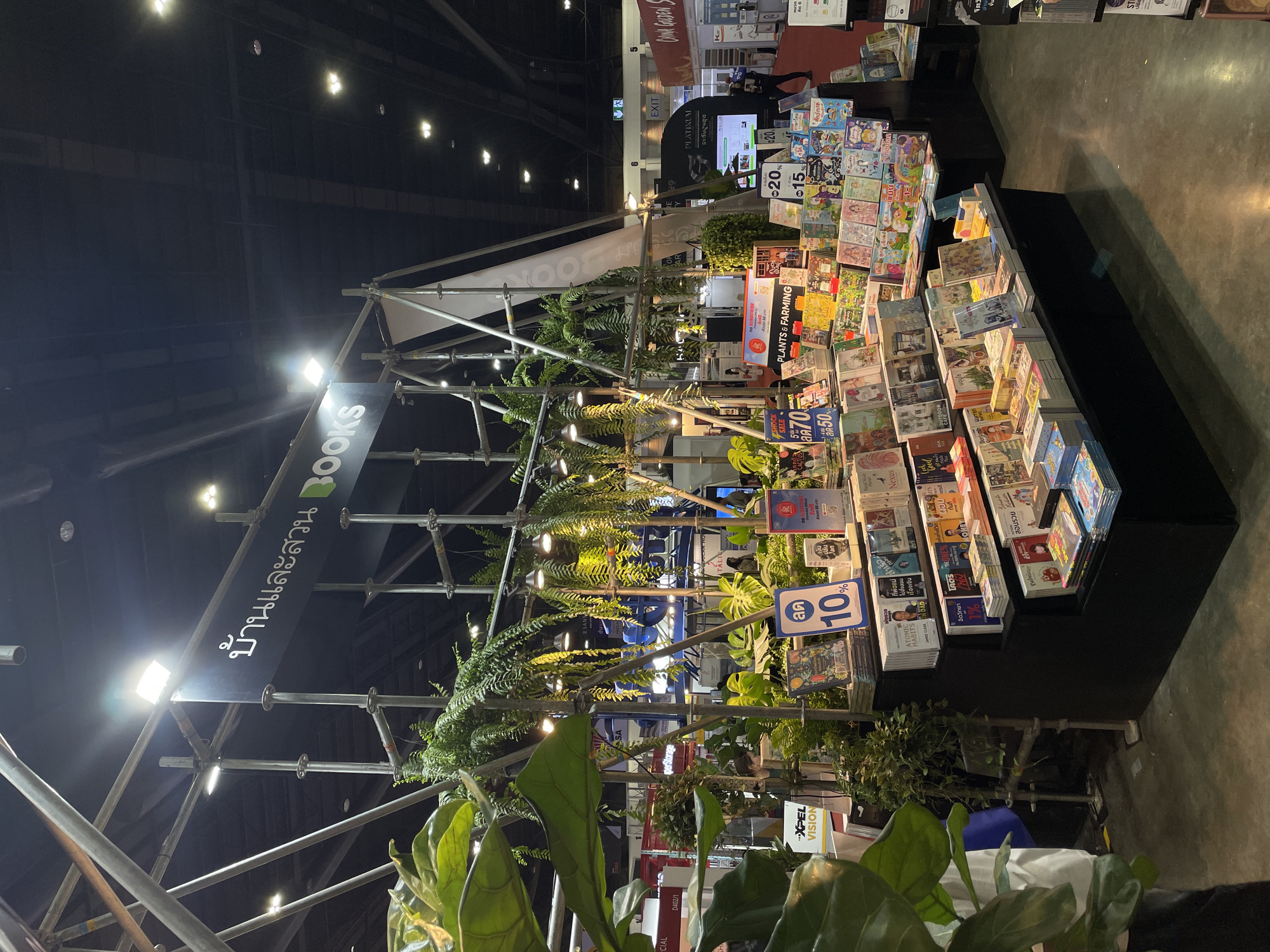 Here's a description of the image:
The image shows a book stall at what appears to be a book fair or exhibition. The location is inferred to be Pak Kret City Municipality, Thailand based on text in the image. The stall is constructed with a metal scaffolding frame, decorated with greenery. A sign with Thai script and the word "BOOKS" is prominently displayed. The stall is filled with books displayed at varying angles. Some books have price tags or promotional stickers on them, with discounts like "10% off" and sale prices in Thai Baht like "50," "70". A sign for "PLANTS & FARMING" can also be seen. The aisle beside the stall is dark, though some people are visible in the background. Additional signage with "EXIT," "PLATINUM" is also visible. The lighting is bright, with spotlights directed at the stall and its contents. The overall impression is of a bustling book fair or exhibition.